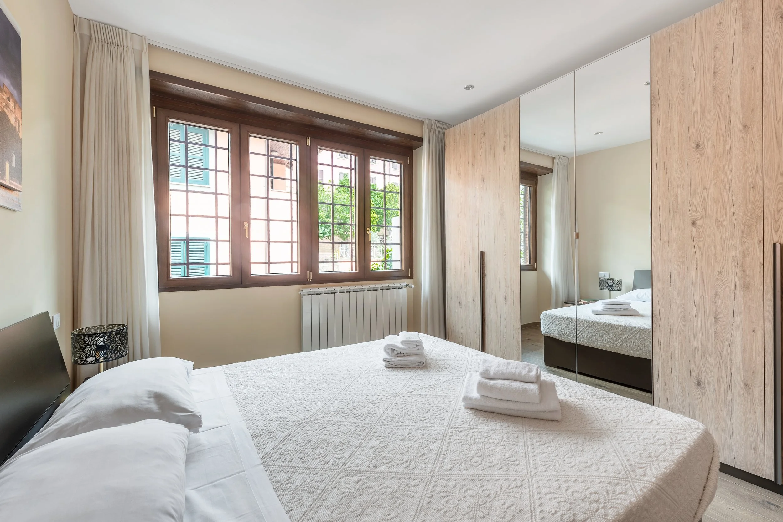 A tidy bedroom with a large window and wooden frame, a bed with folded towels on top, a mirrored closet door, and a small bedside table with a lamp.