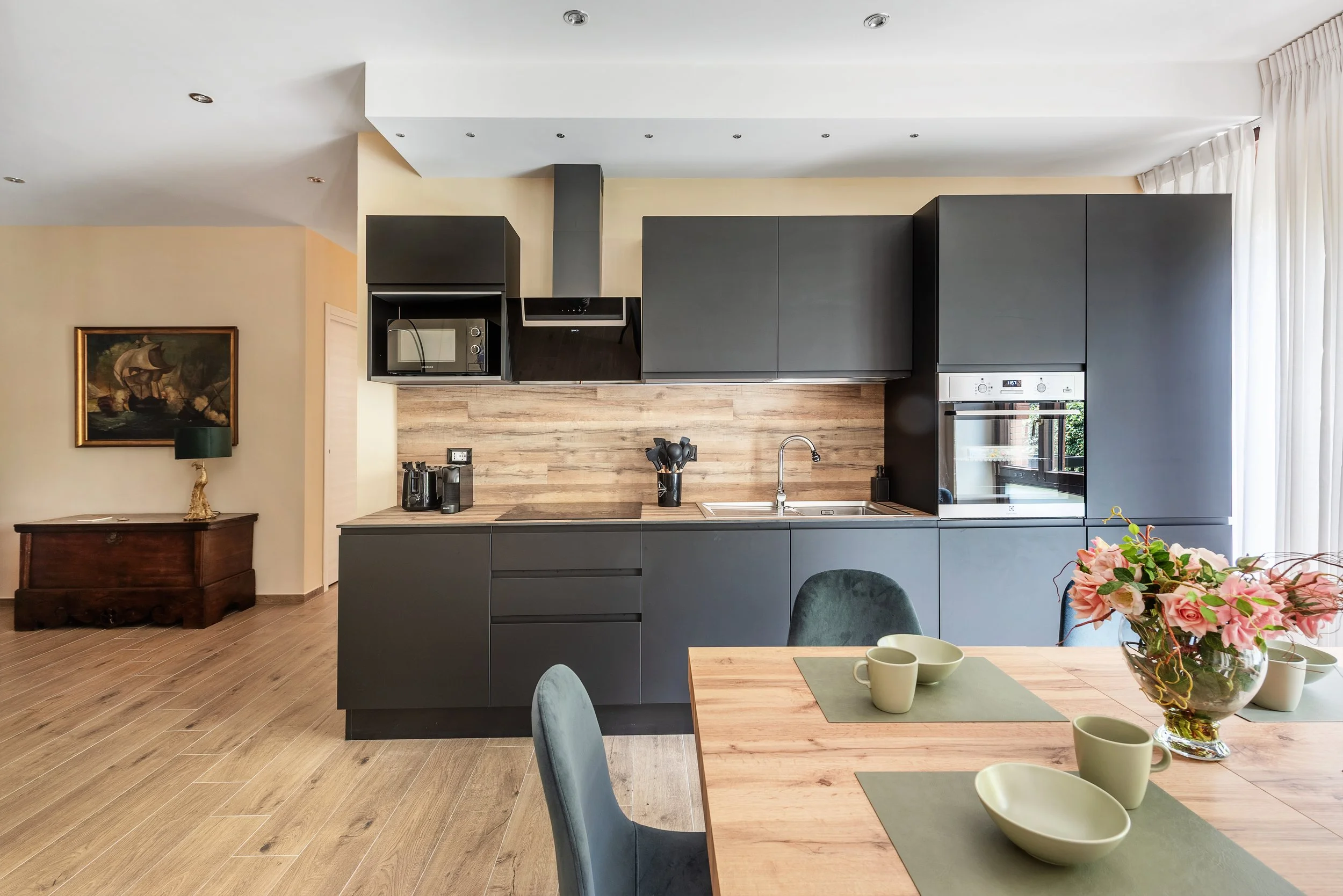 Modern kitchen with black cabinets, wooden backsplash, stainless steel appliances, and a dining table with a floral centerpiece.