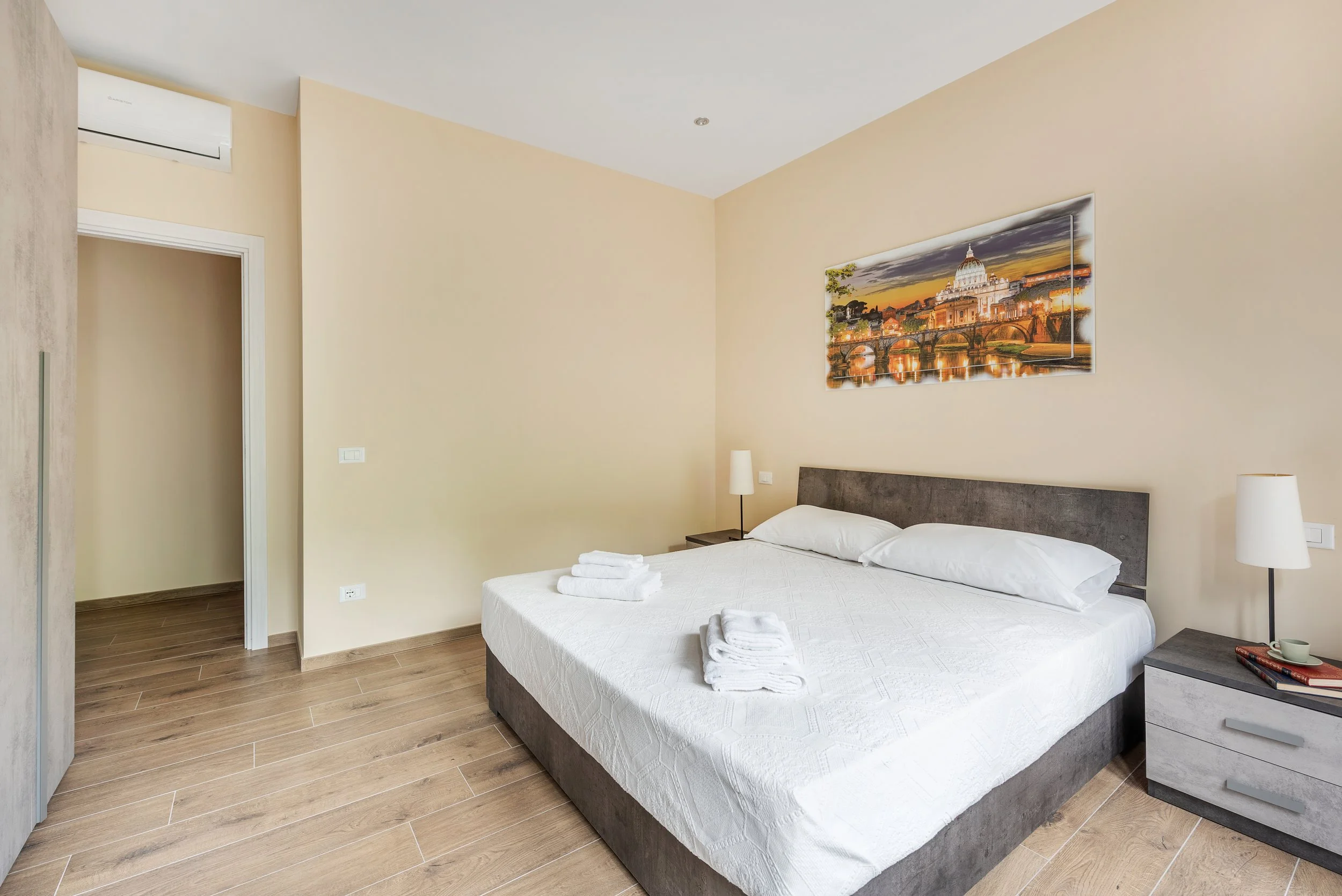 Comfortable bedroom with a king-sized bed, two bedside tables with lamps, framed cityscape art on cream walls, and a wooden floor.
