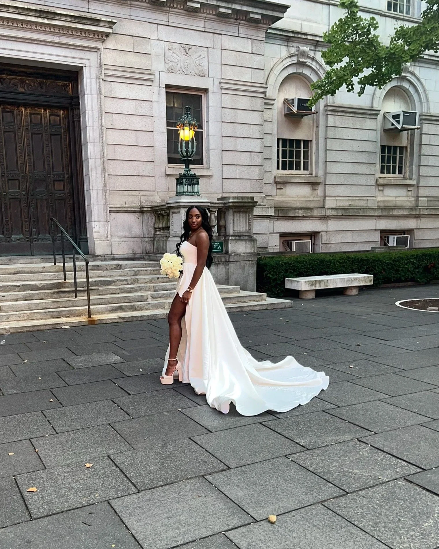 A woman in a white wedding gown holding a bouquet of white roses, standing on a city sidewalk in front of an ornate building with stone steps and a decorative door.