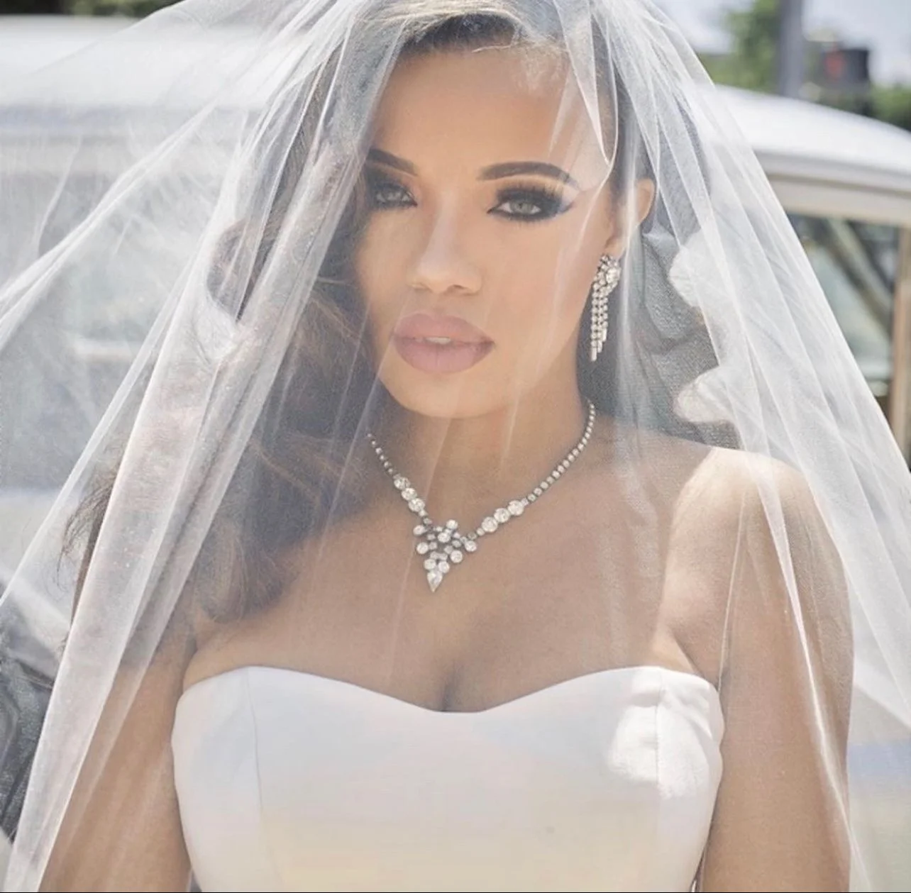 A bride wearing a white strapless wedding dress, veil, and pearl jewelry, with makeup and hairstyle, outdoors.