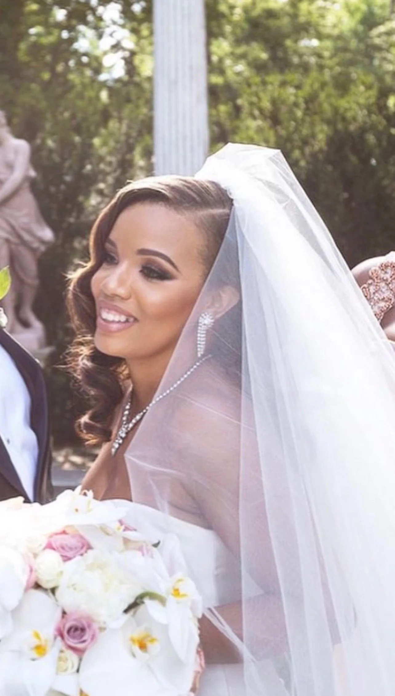 Bride smiling with a white veil, holding a bouquet of white and pink roses, outdoors with sunlight filtering through trees.