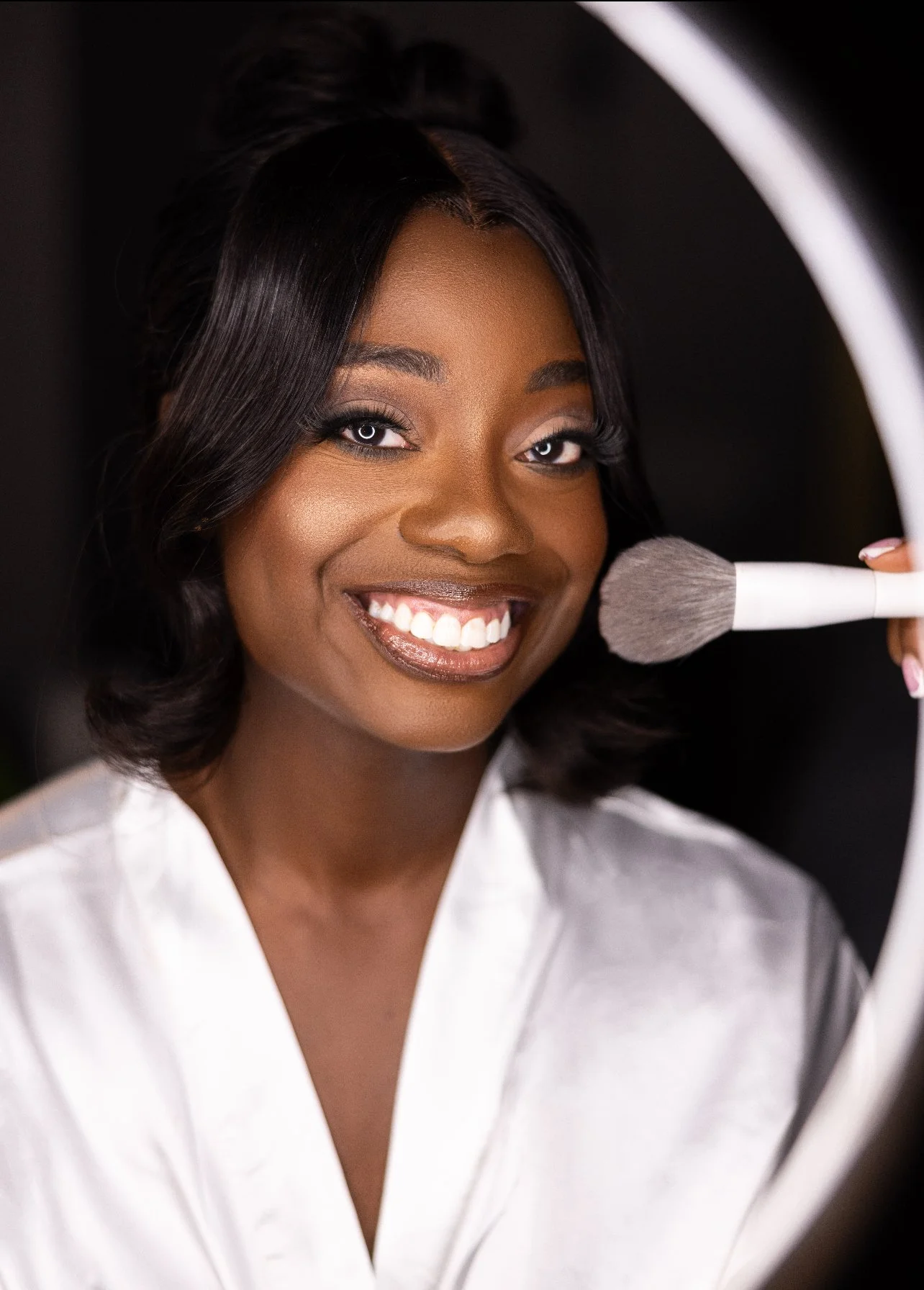 Woman with dark hair smiling and holding makeup brush near her face, in front of a mirror.