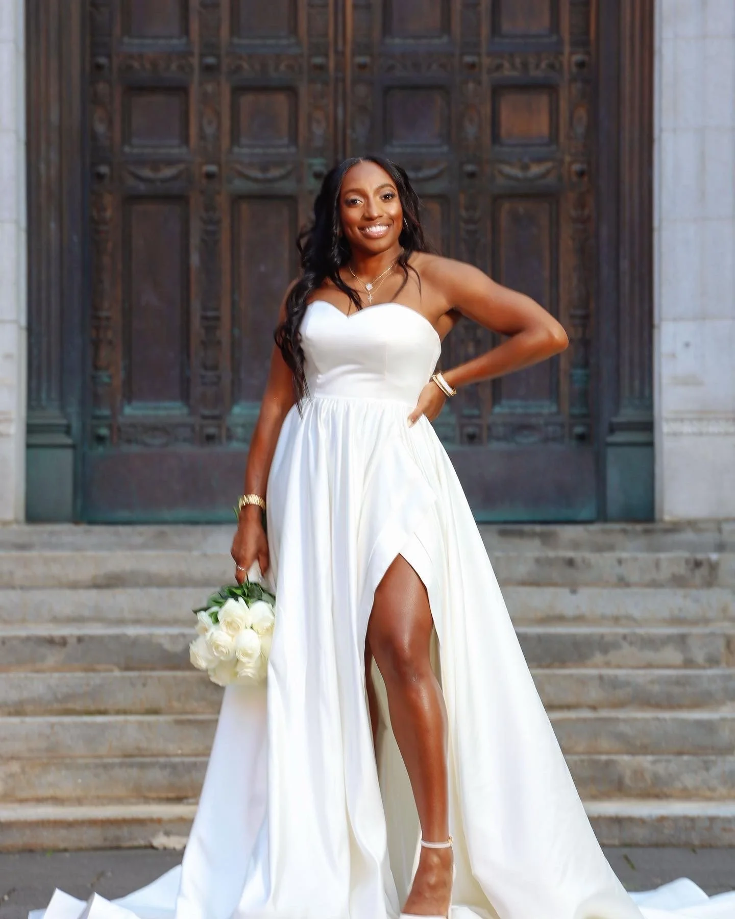 A woman in a white dress holding a bouquet of white roses, standing on steps in front of a wooden door.