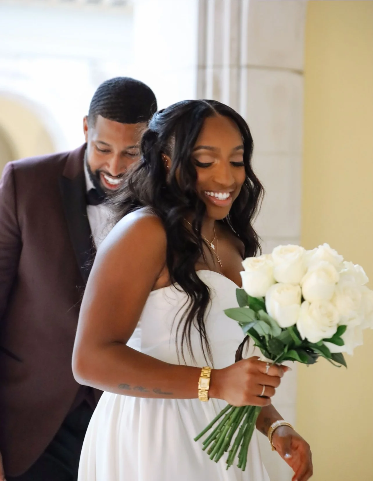 A smiling bride holding a bouquet of white roses and a groom behind her, both dressed in formal wedding attire, inside a bright indoor venue.