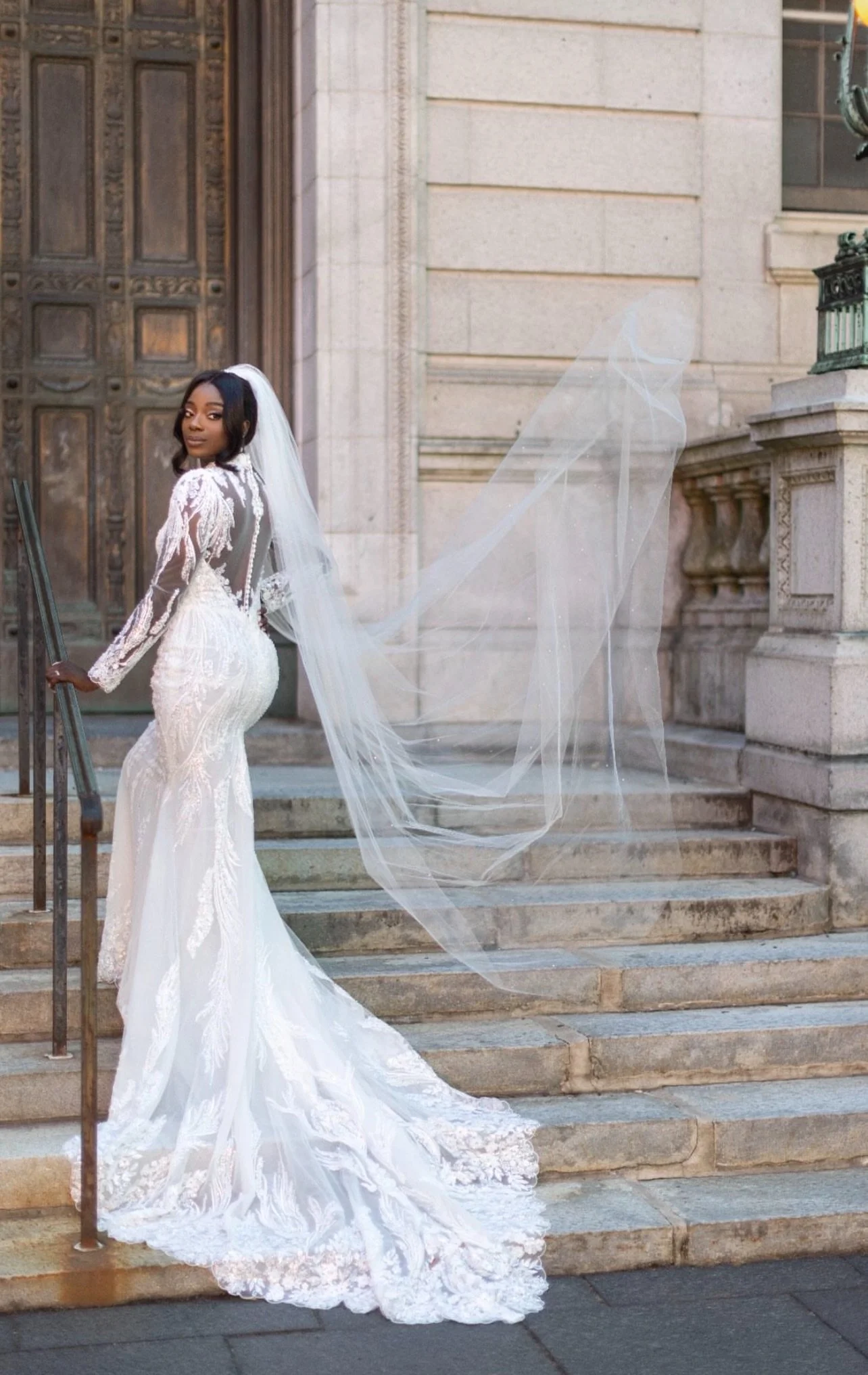 A bride in a white wedding gown and veil standing on stone steps in front of a historic building with large wooden doors.