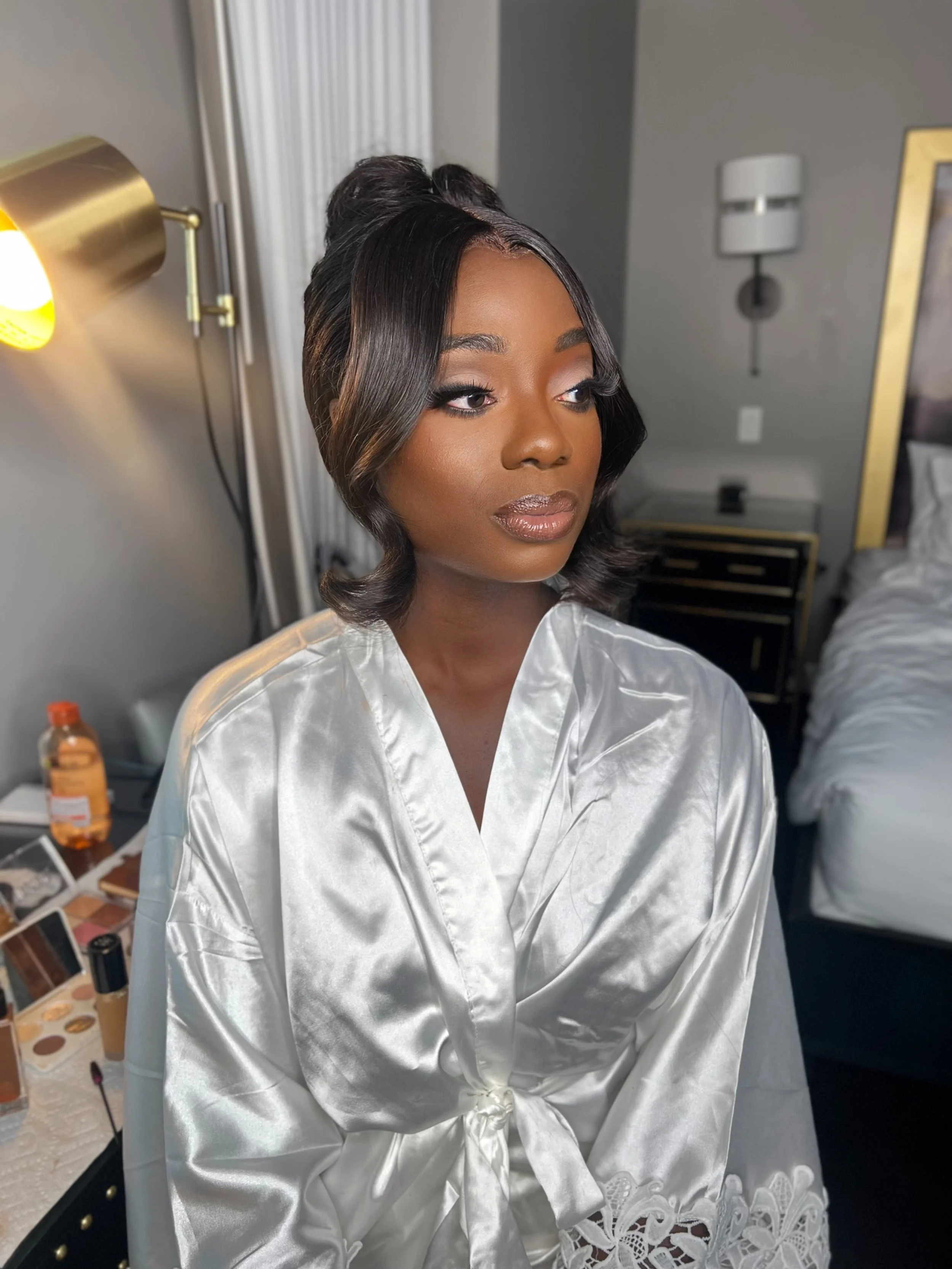 A woman with styled hair and makeup in a satin robe, sitting in a hotel room.