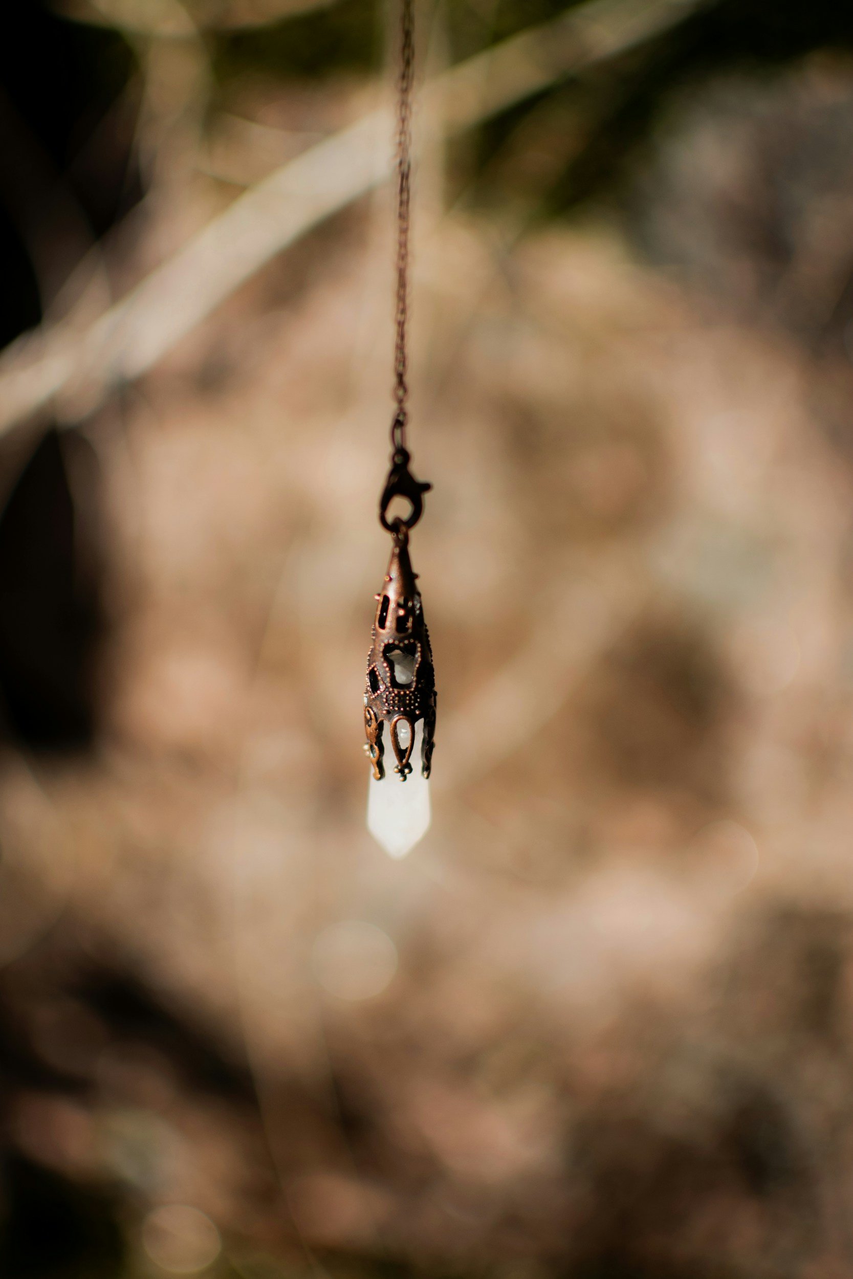 A close-up of a vintage necklace with a hanging pendant, featuring an elongated, pointed crystal, suspended against a blurred natural background.