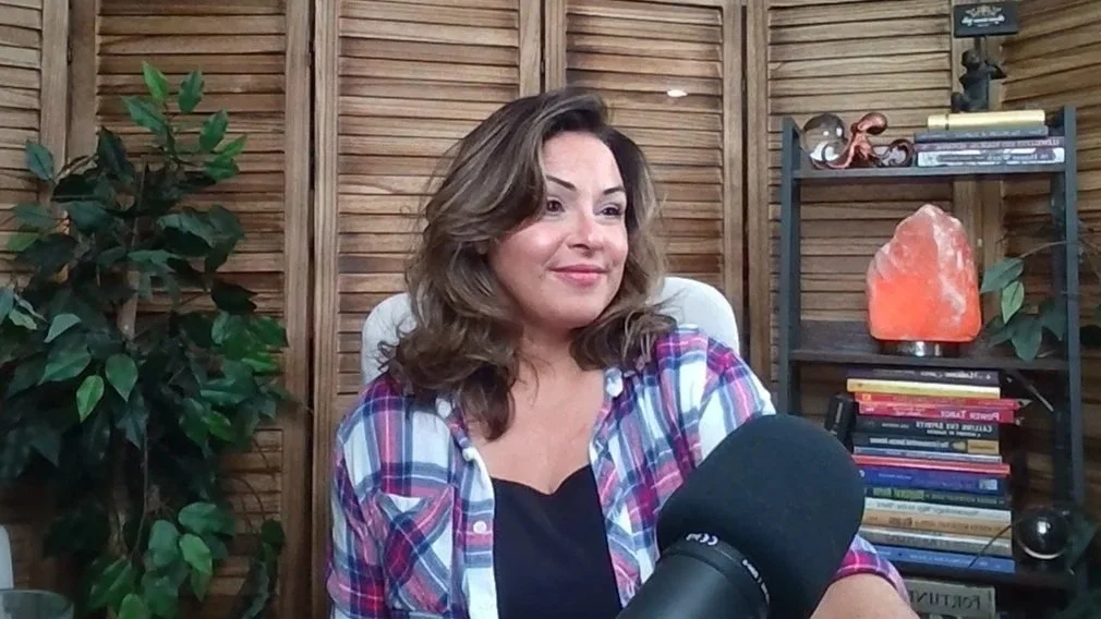 A woman with shoulder-length brown hair wearing a plaid shirt and black top sits in front of a microphone in a room with wooden walls. There are plants and a bookshelf with books and decorative items behind her.