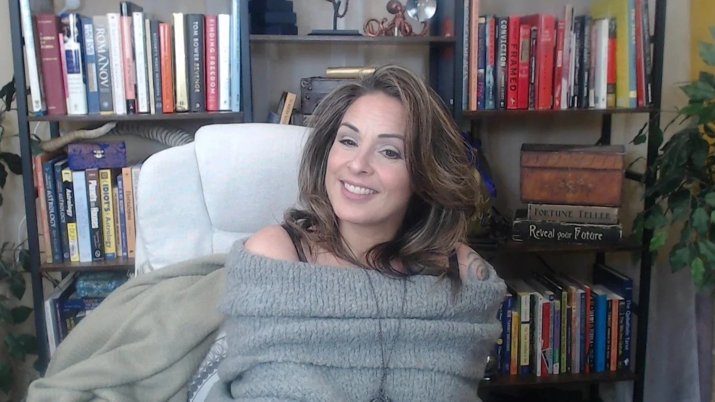 A woman with shoulder-length brown hair smiling and sitting in a white office chair in front of a bookshelf filled with books and decorative items.