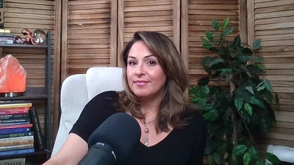 A woman with shoulder-length brown hair and a black shirt sitting in a white chair in front of a wooden paneled wall, with a bookshelf and a large green plant behind her.