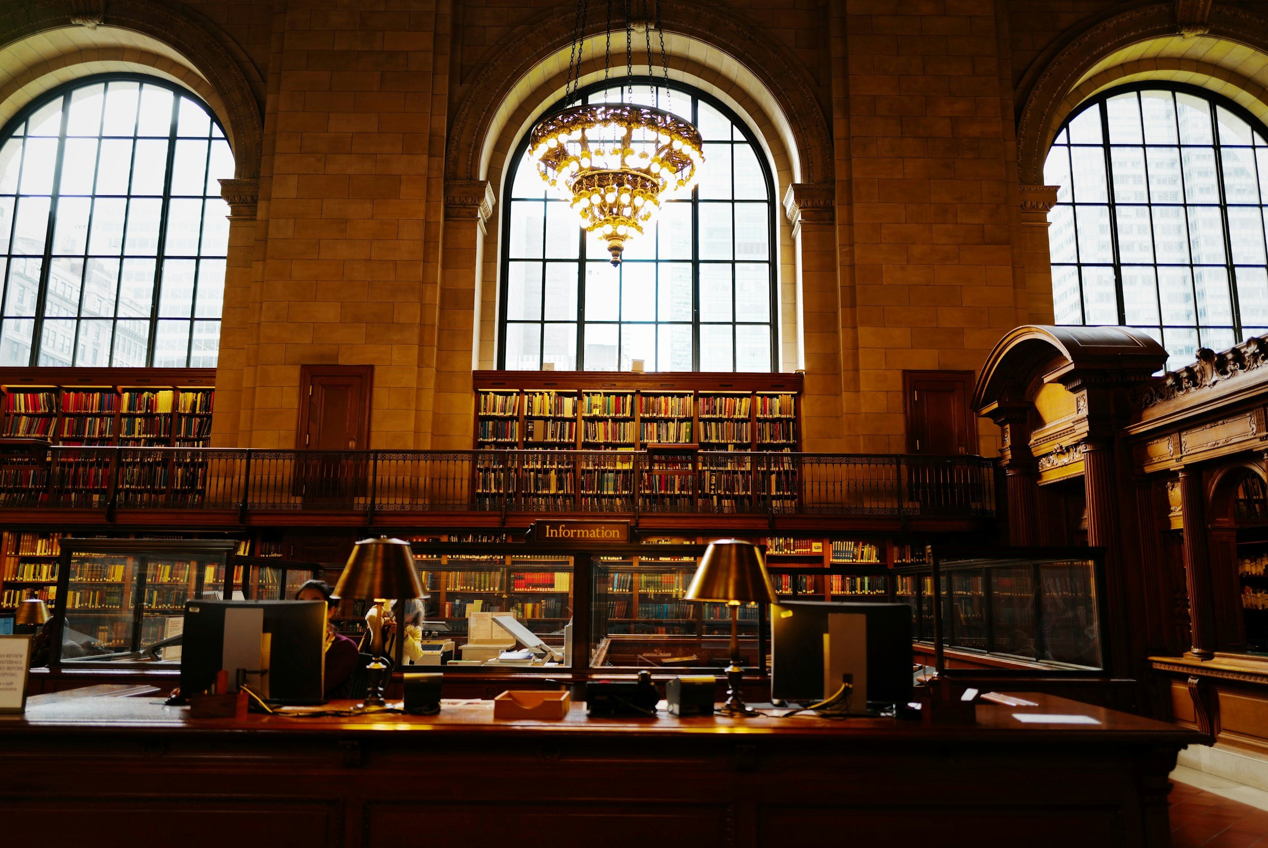 Interior of a grand library with large windows, a chandelier, and rows of bookshelves behind a wooden information desk.