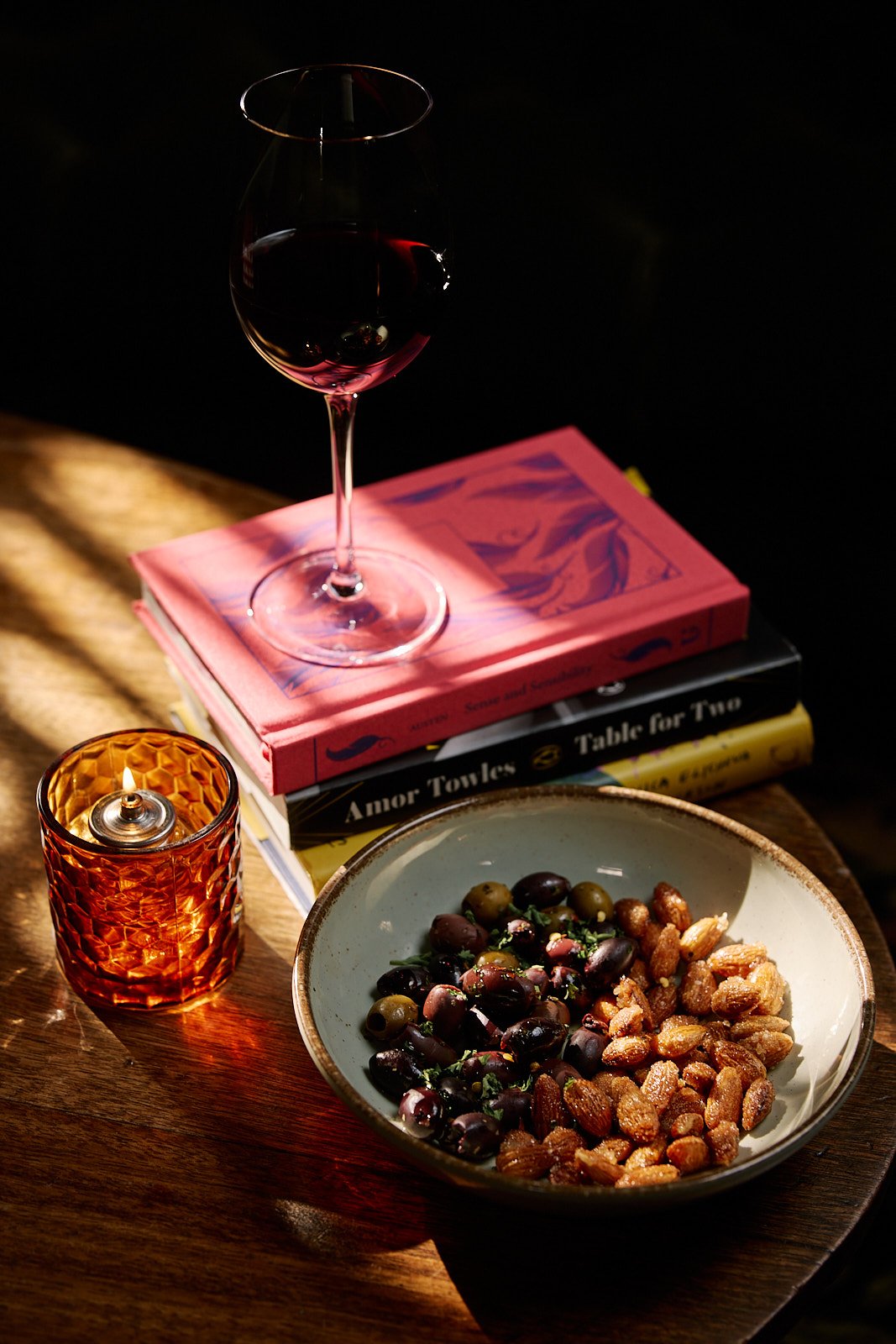 bowl of almonds and olives next to a stack of books with a wine glass sitting on top and a candle