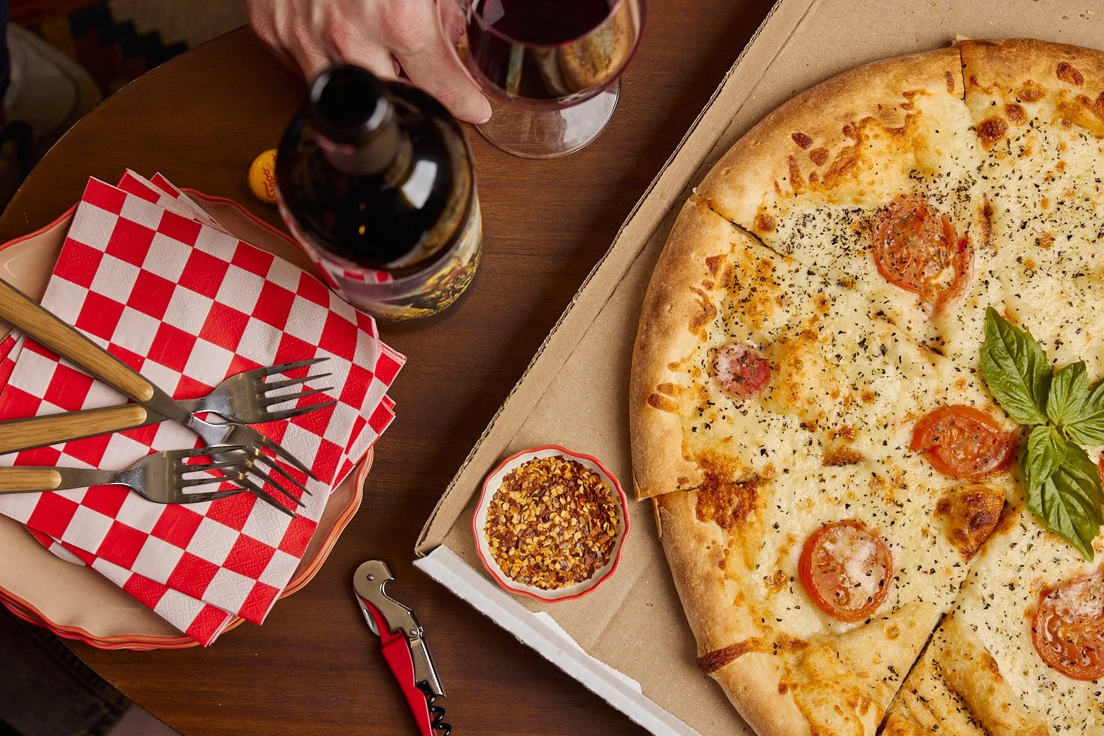 scene of a table with a pizza box, red checkered napkins, and a wine bottle.