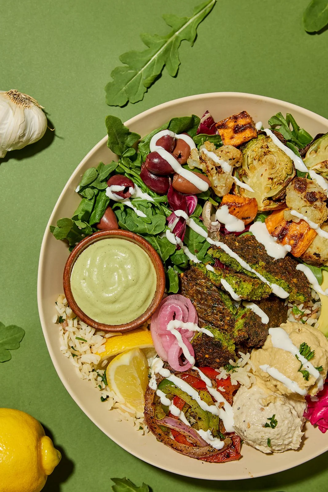 bowl of mediteranean food with falafel, olives, arugula, and green tahini sauce, styled on a green solid colored backdrop with ingredients around it.