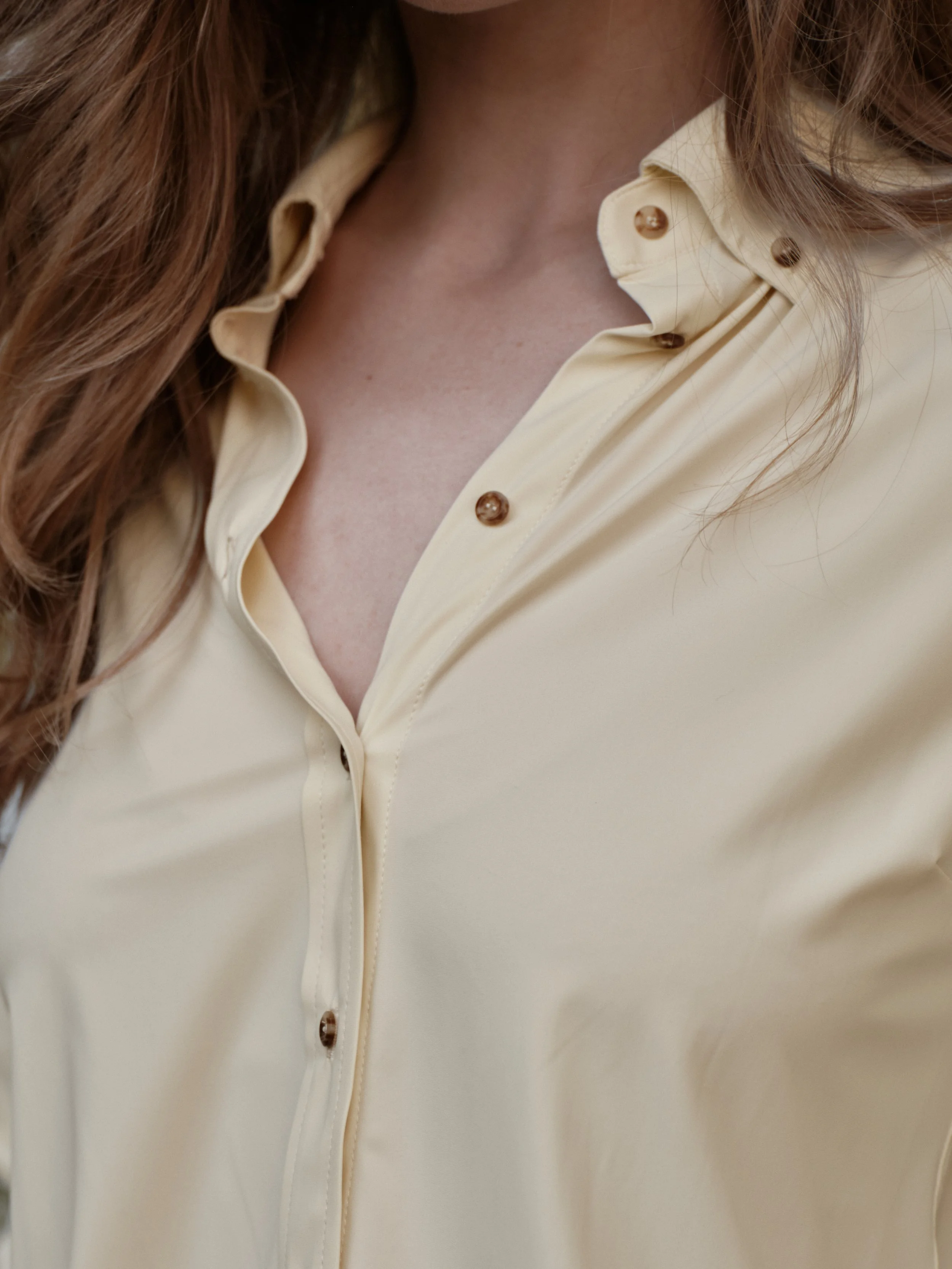 Close-up of a woman's chest and neck, showing part of a light beige button-up shirt with brown buttons, and brown wavy hair.