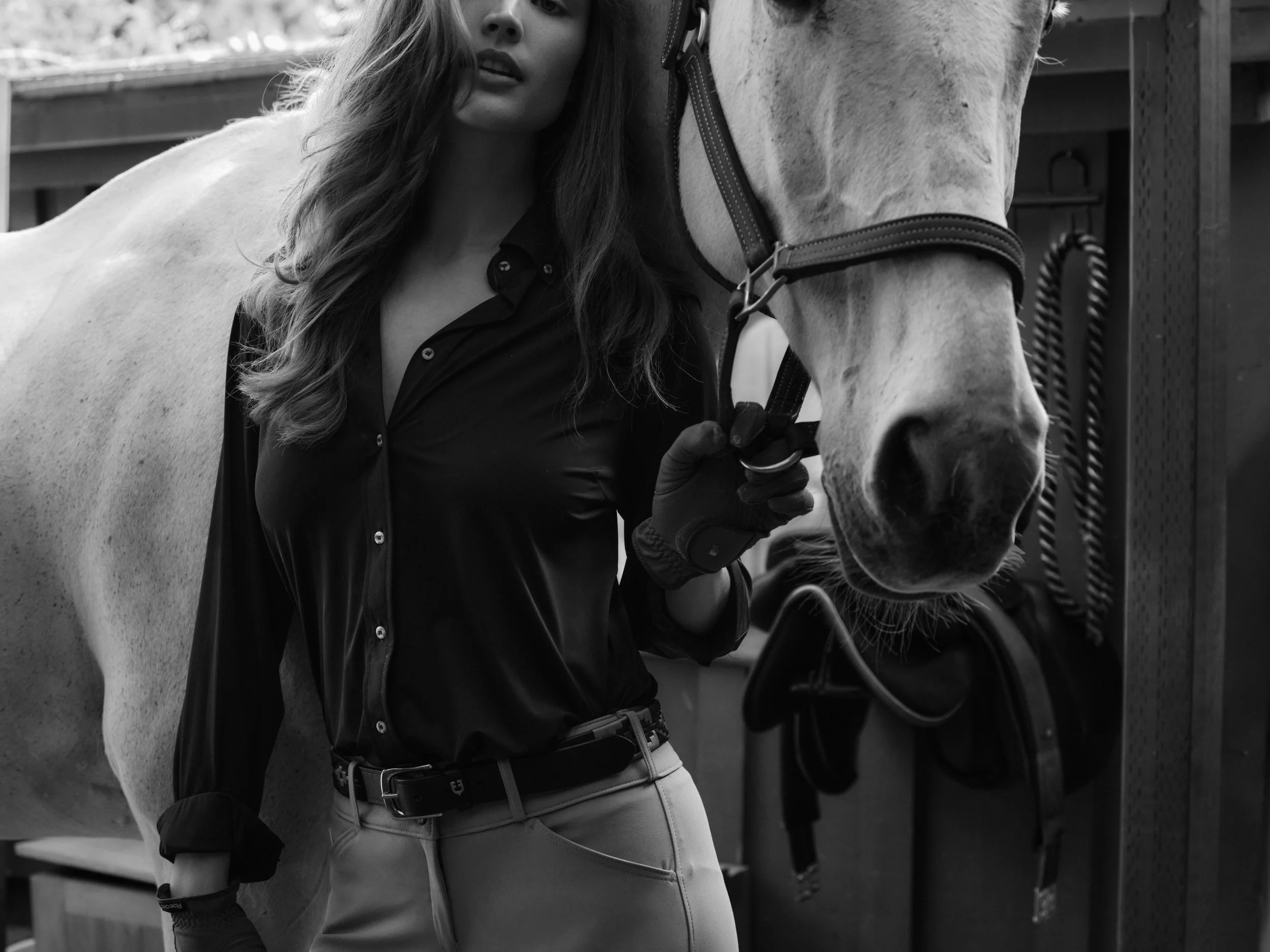 A woman in riding attire holding the reins of a white horse, in an outdoor stable setting.