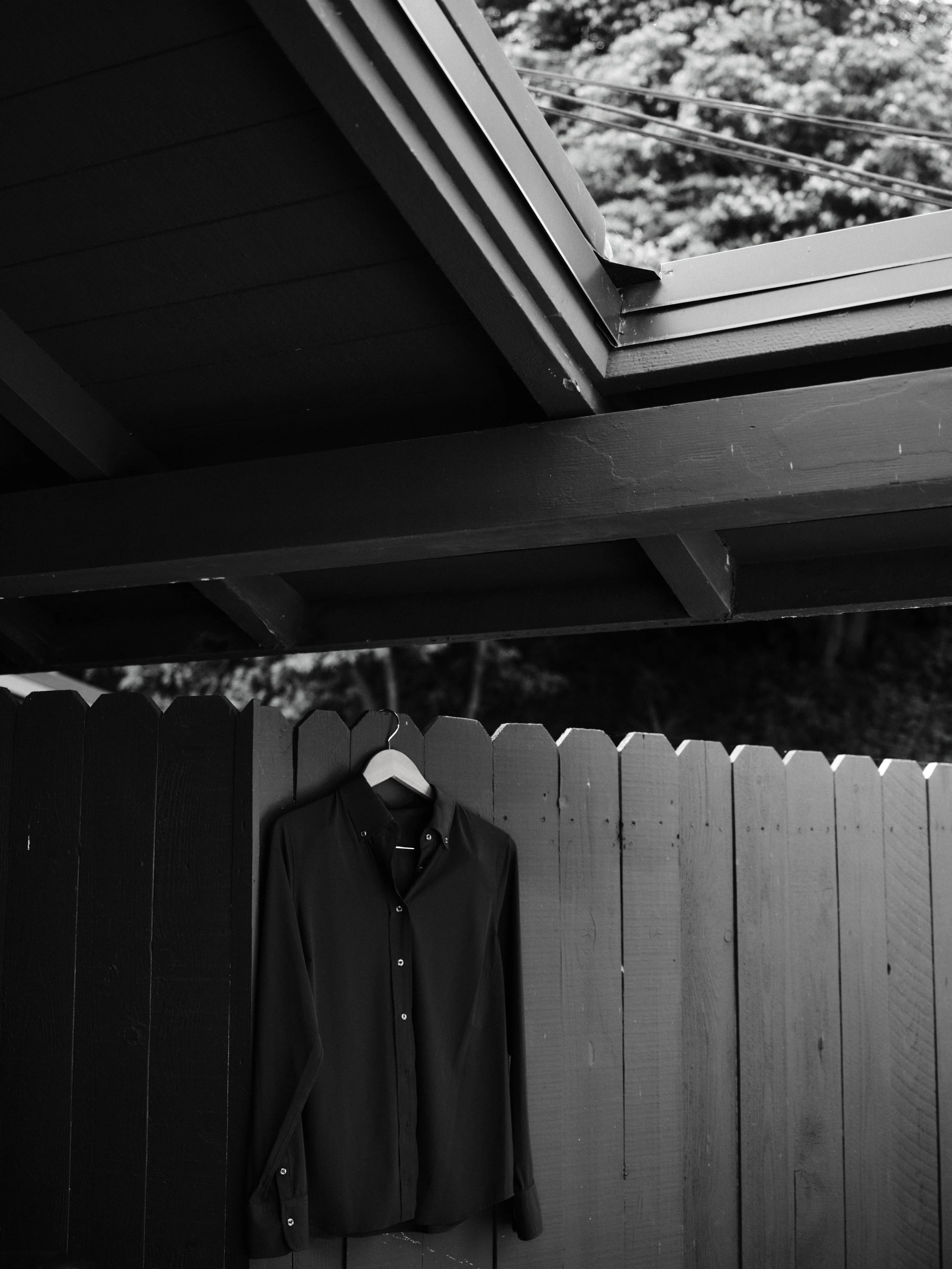 A black shirt hangs from a hook on a dark-colored fence in a backyard. Behind the fence, there are trees and power lines visible.