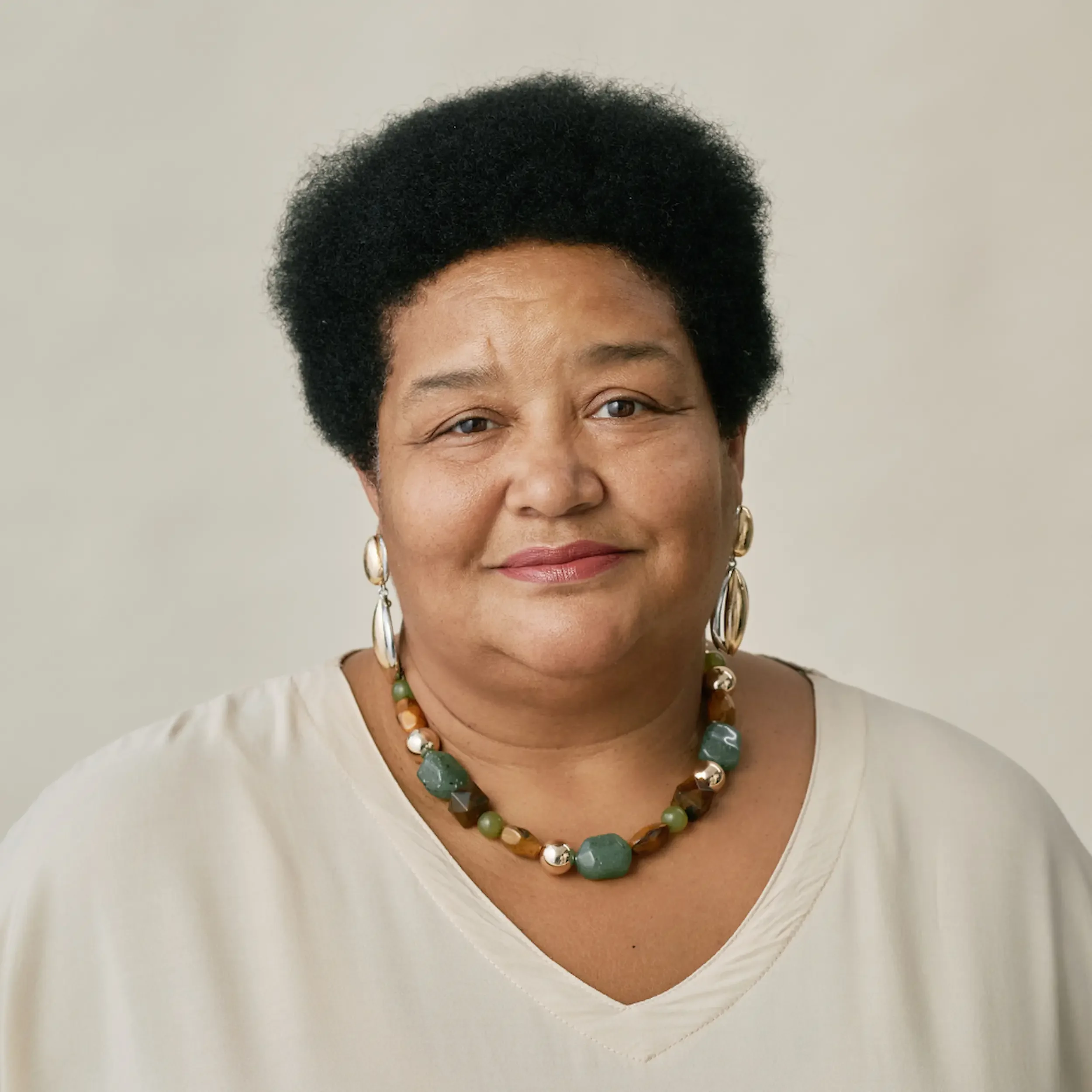A middle-aged woman with short black hair smiling, wearing a beige blouse, colorful beaded necklace, and hoop earrings, against a plain light-colored background.