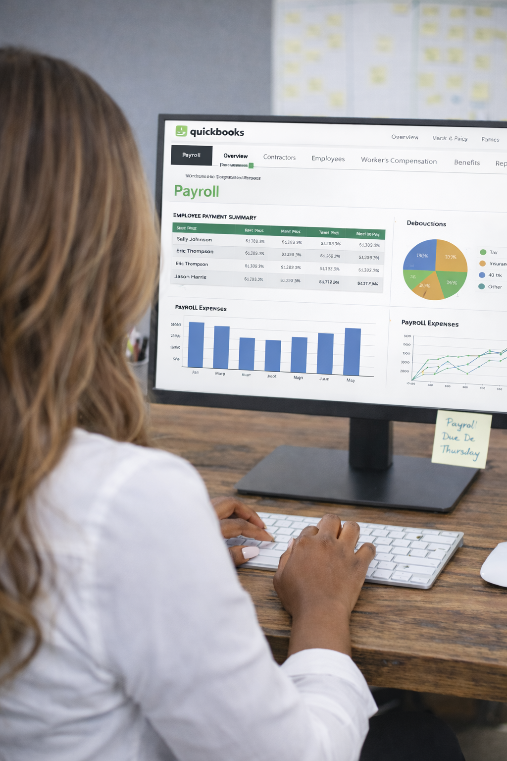 A person working at a desk on a computer displaying a QuickBooks payroll dashboard, with charts and employee payment information visible on the screen.