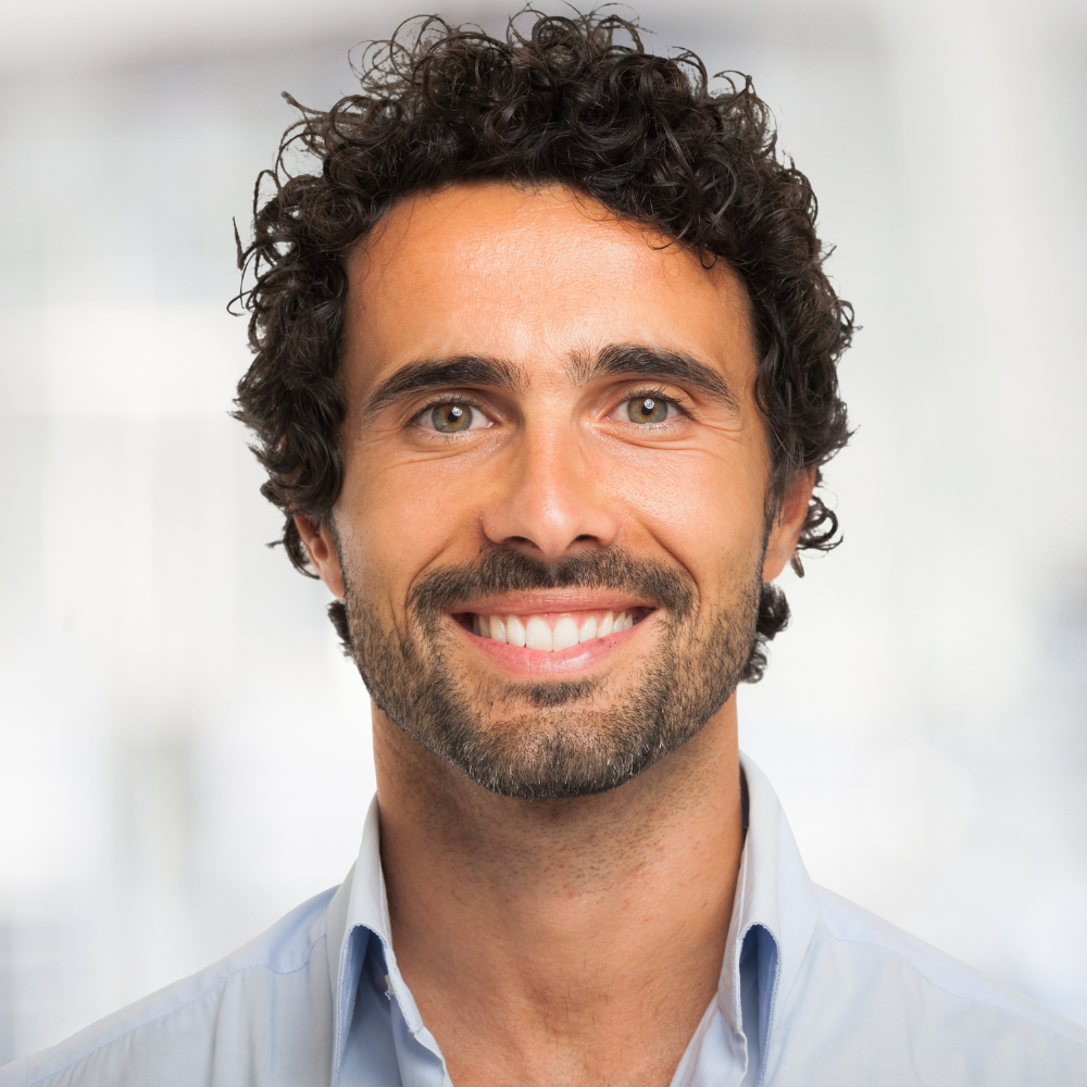 Headshot of a man with curly dark hair, light skin tone, and a beard, smiling in a professional setting.