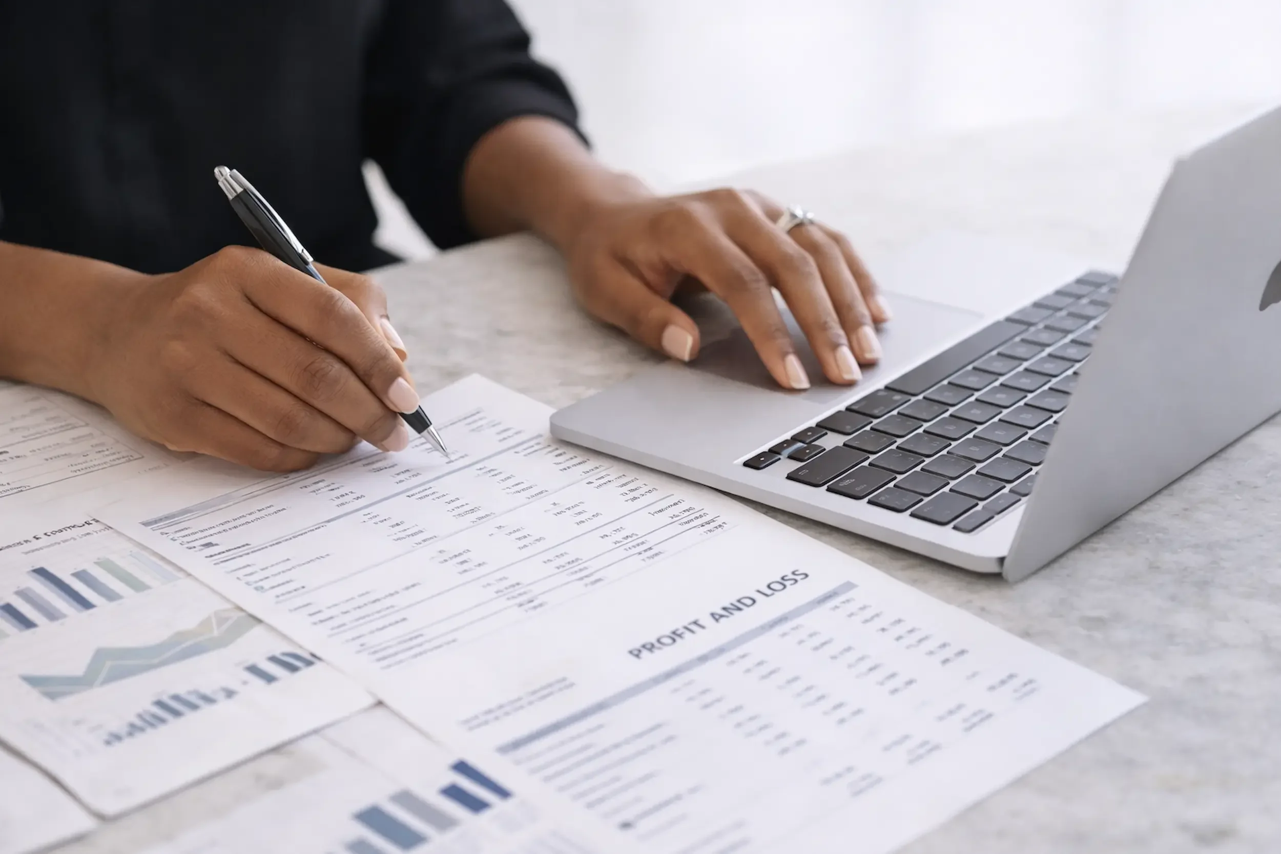 Person using a laptop computer with financial documents and papers with graphs and charts on a desk.