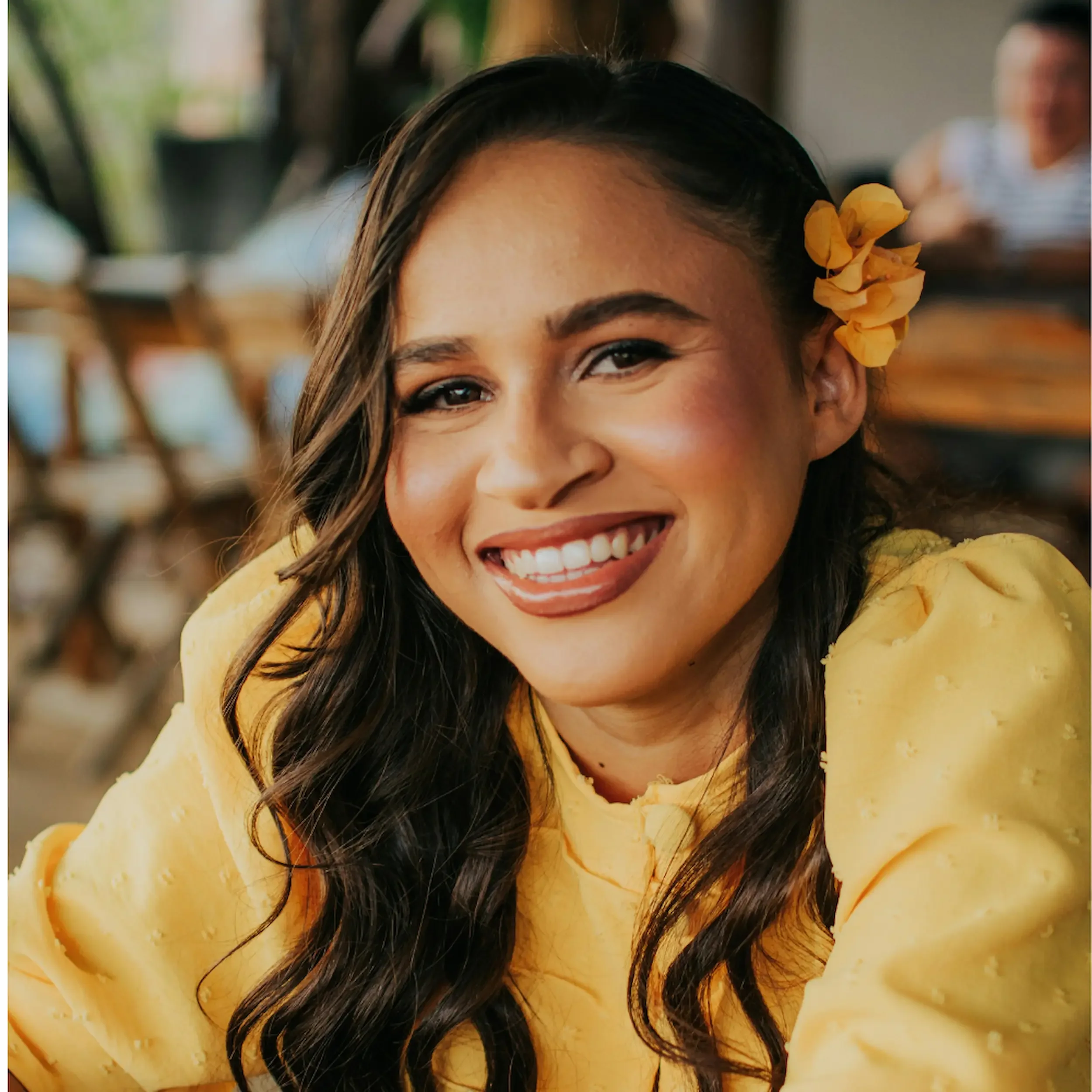 A woman smiling with long dark hair, wearing a yellow top and a yellow flower in her hair, indoors with a blurred background.