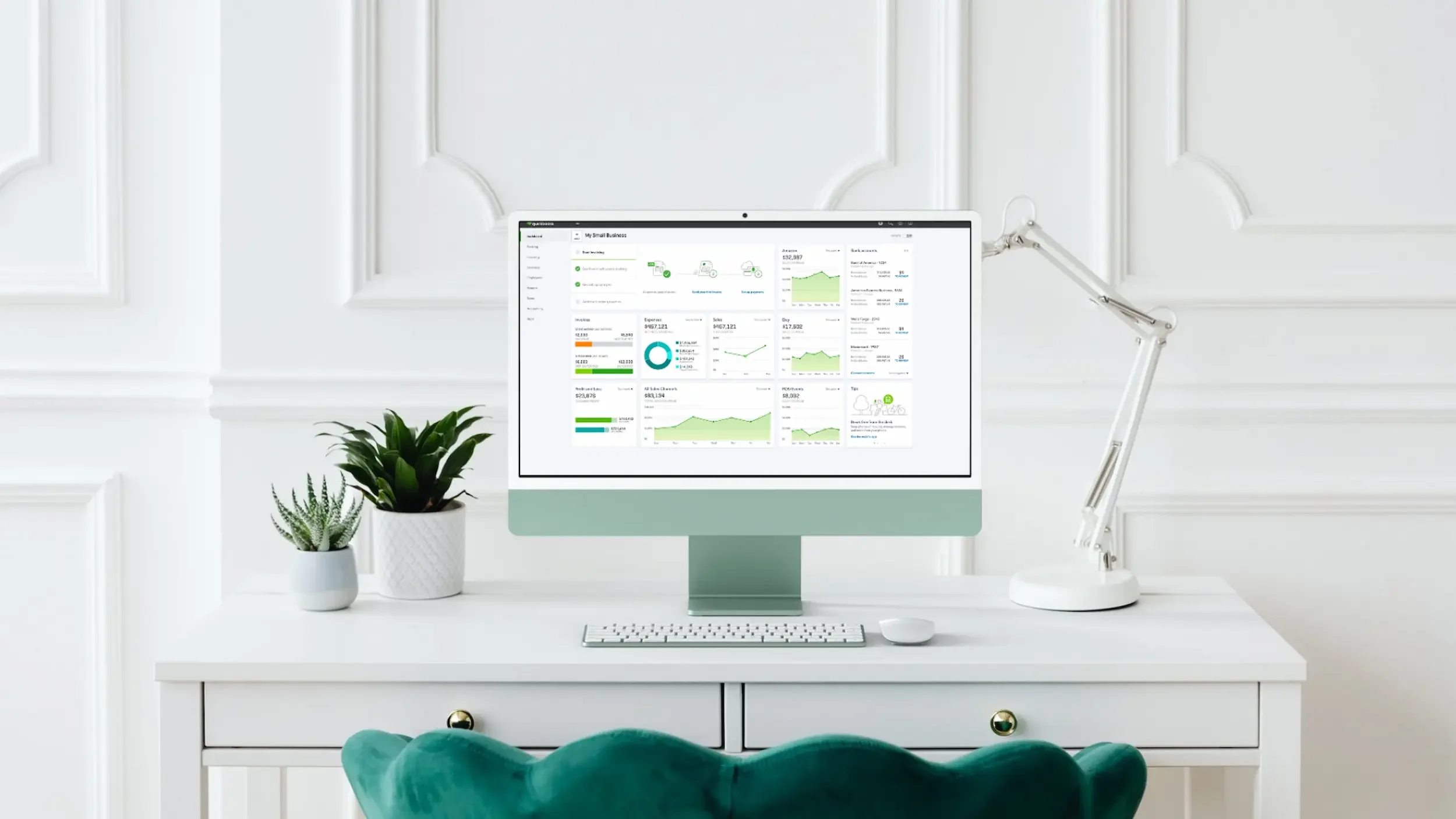 A white desk with two potted plants, one small and one medium, beside a computer monitor displaying business analytics, a wireless keyboard and mouse, and a modern white desk lamp in a minimalistic, white walled room with decorative wall molding.
