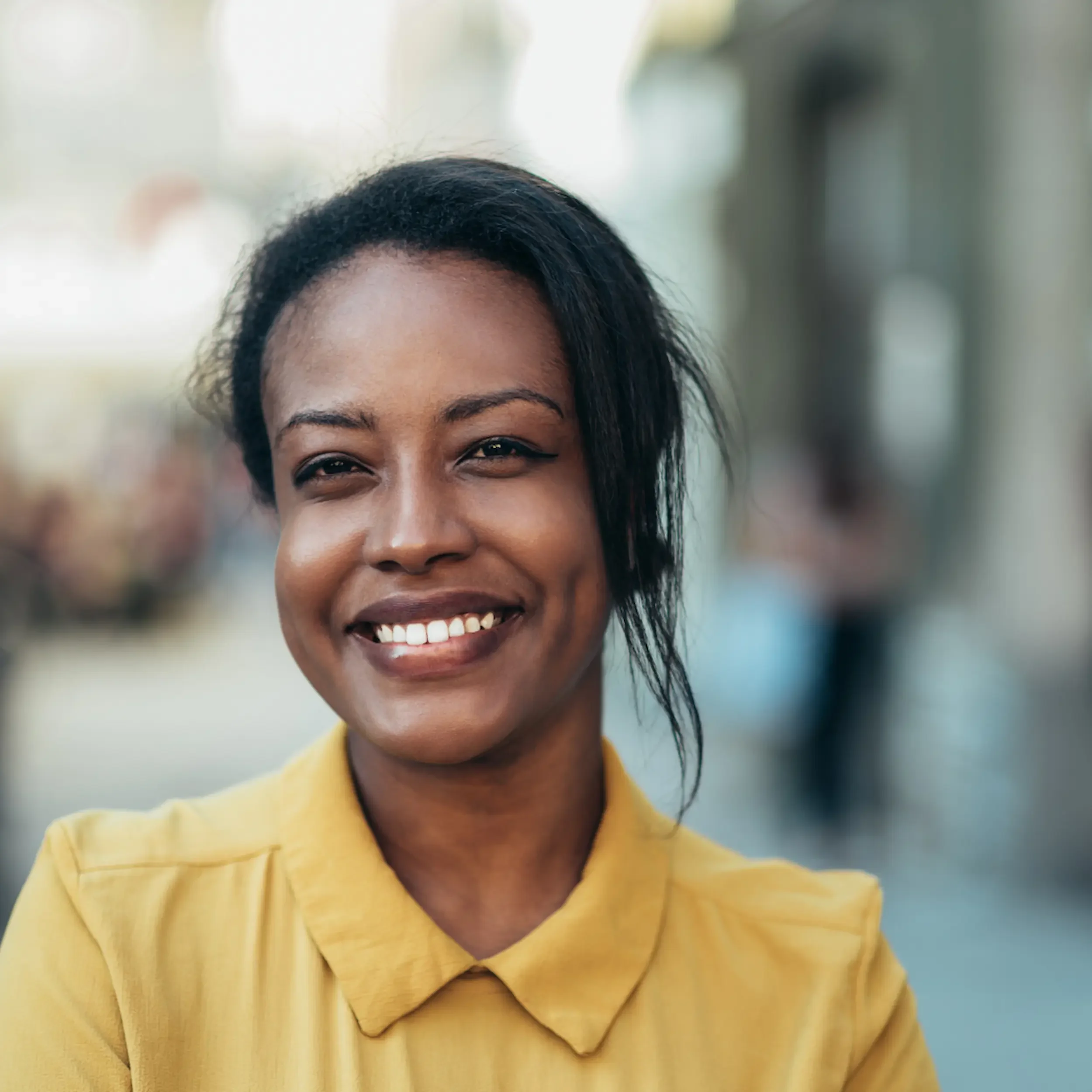A woman smiling outdoors, wearing a yellow shirt.