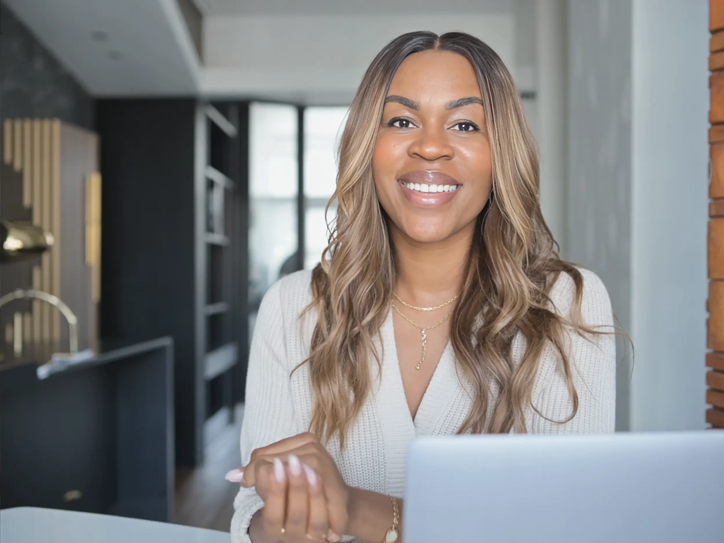 A woman with long wavy hair smiling while working on a laptop in a modern kitchen with black shelves and wooden accents.
