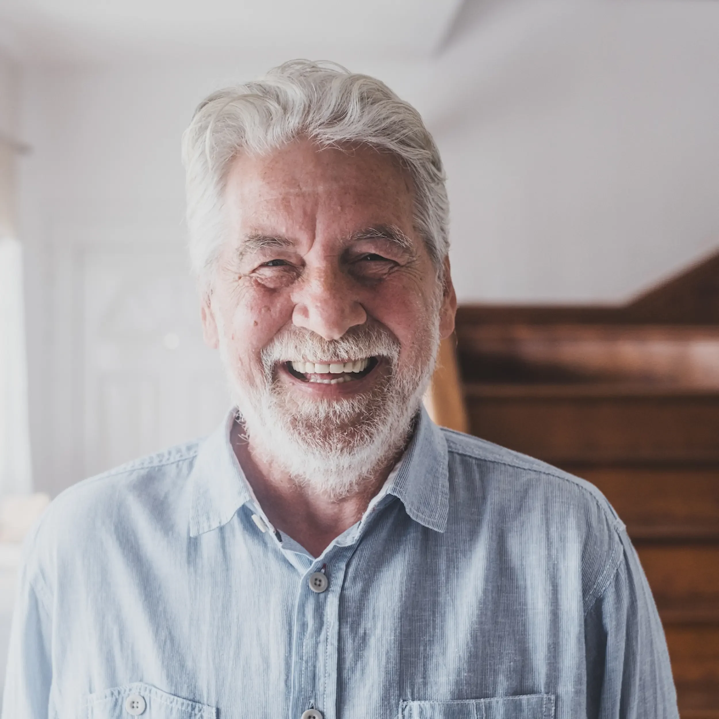 A cheerful elderly man with white hair and a beard, smiling widely in a bright room.