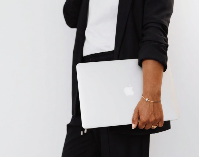 Person wearing a black blazer holding a closed silver MacBook with an Apple logo, standing against a white background.