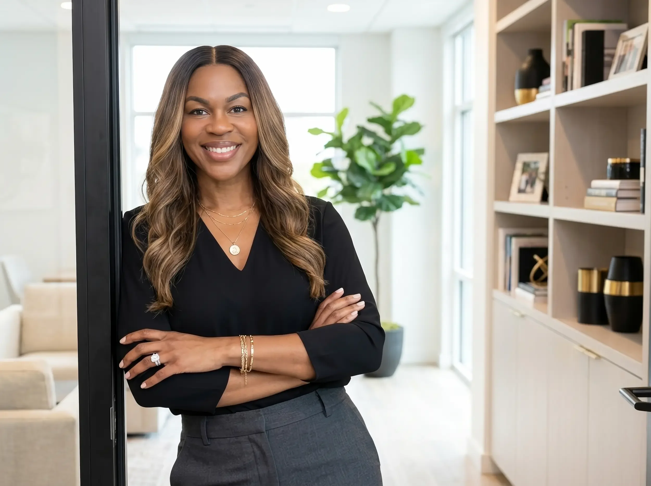 A smiling woman with long wavy hair, wearing a black blazer, gold jewelry, and a ring, standing indoors in front of a bookshelf and a large window.