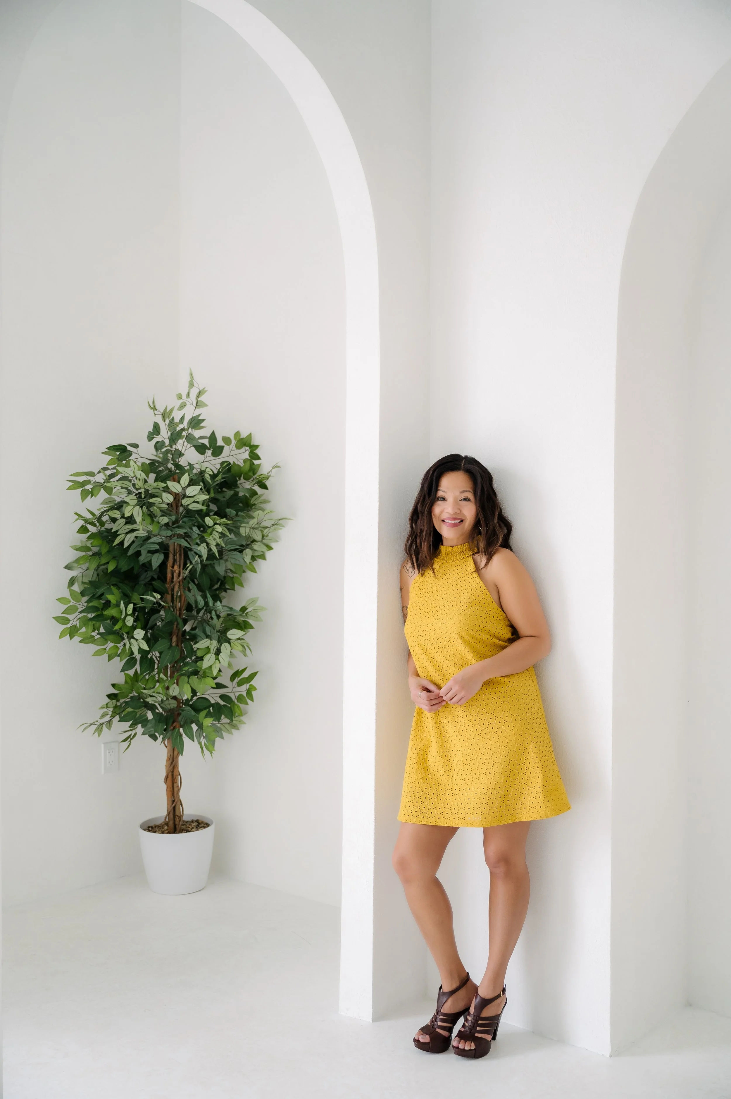 A woman in a yellow dress and high heels standing and smiling in a bright, white room with an indoor plant in a white pot.