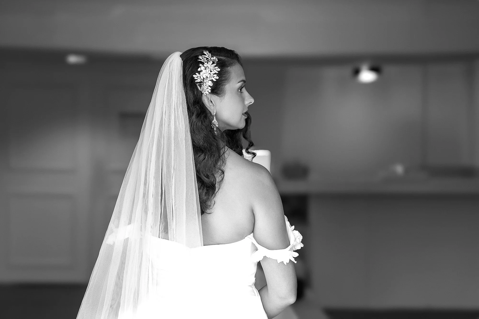 A black and white photograph of a bride from behind, wearing a white strapless gown, a long sheer veil, and a decorative hair clip, looking toward the right side of the frame.