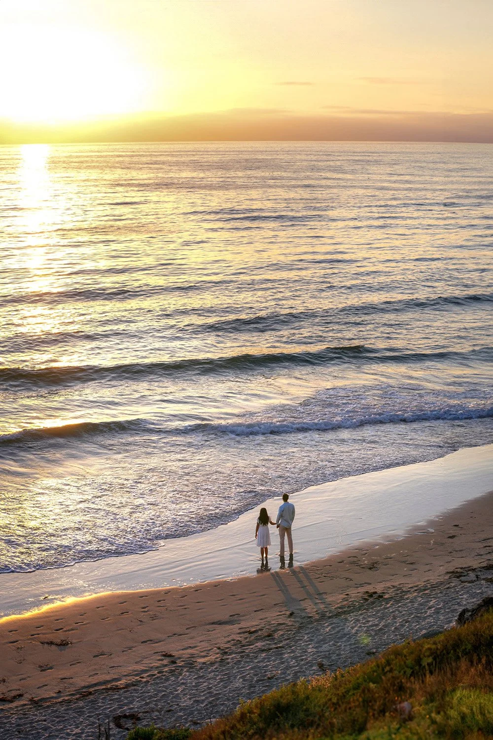 An outdoor photograph of a couple holding hands while standing on a sandy beach at sunset. The couple, viewed from behind, looks out at the ocean as waves roll onto the shore, under a sky with the bright, low sun casting a warm golden light across th