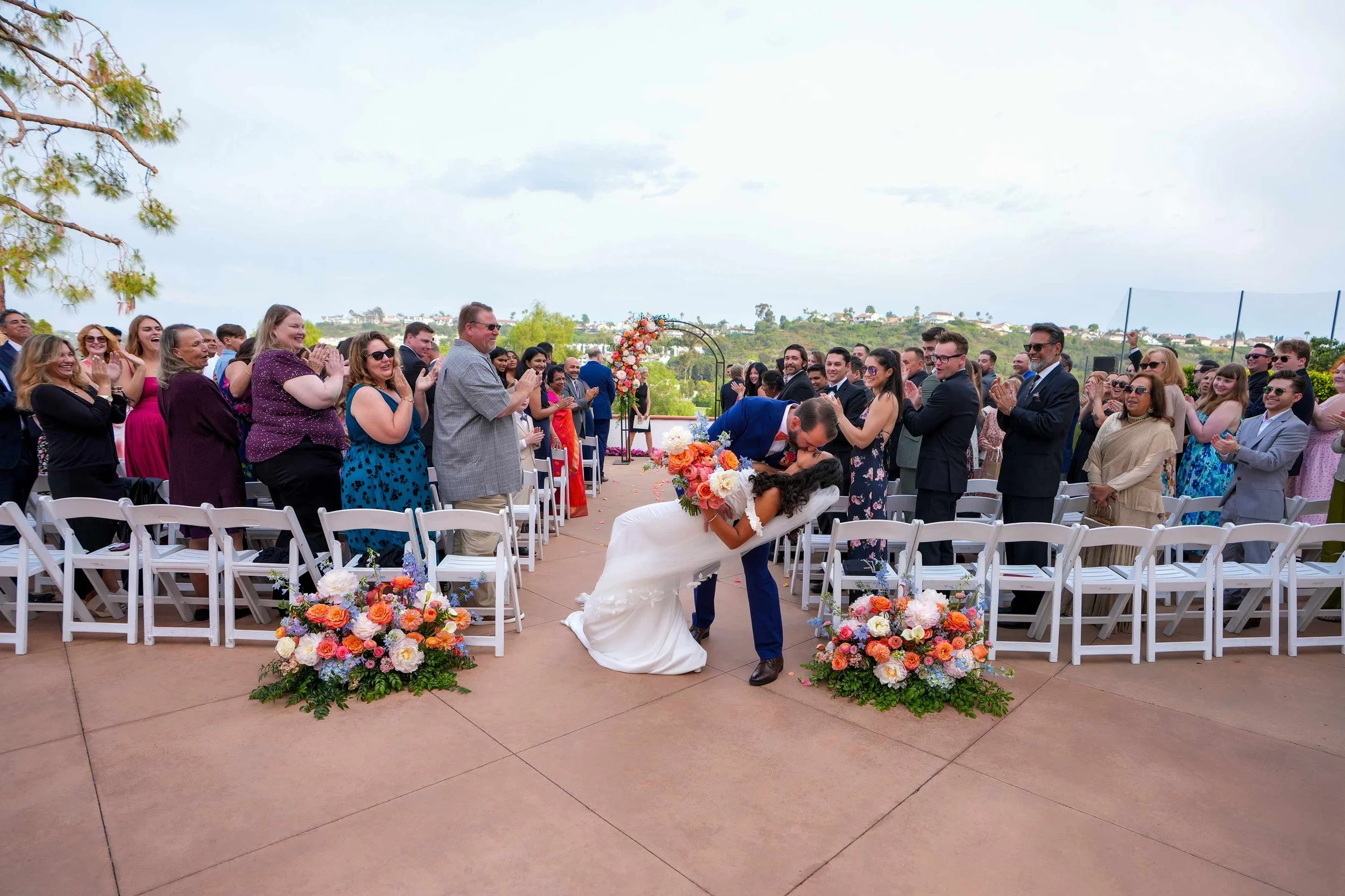 A bride in a white dress and a groom in a blue suit share a kiss in a dramatic dip at the end of an outdoor wedding aisle in Omni Resort, Carlsbad, surrounded by applauding guests in white chairs and vibrant orange and blue floral arrangements.