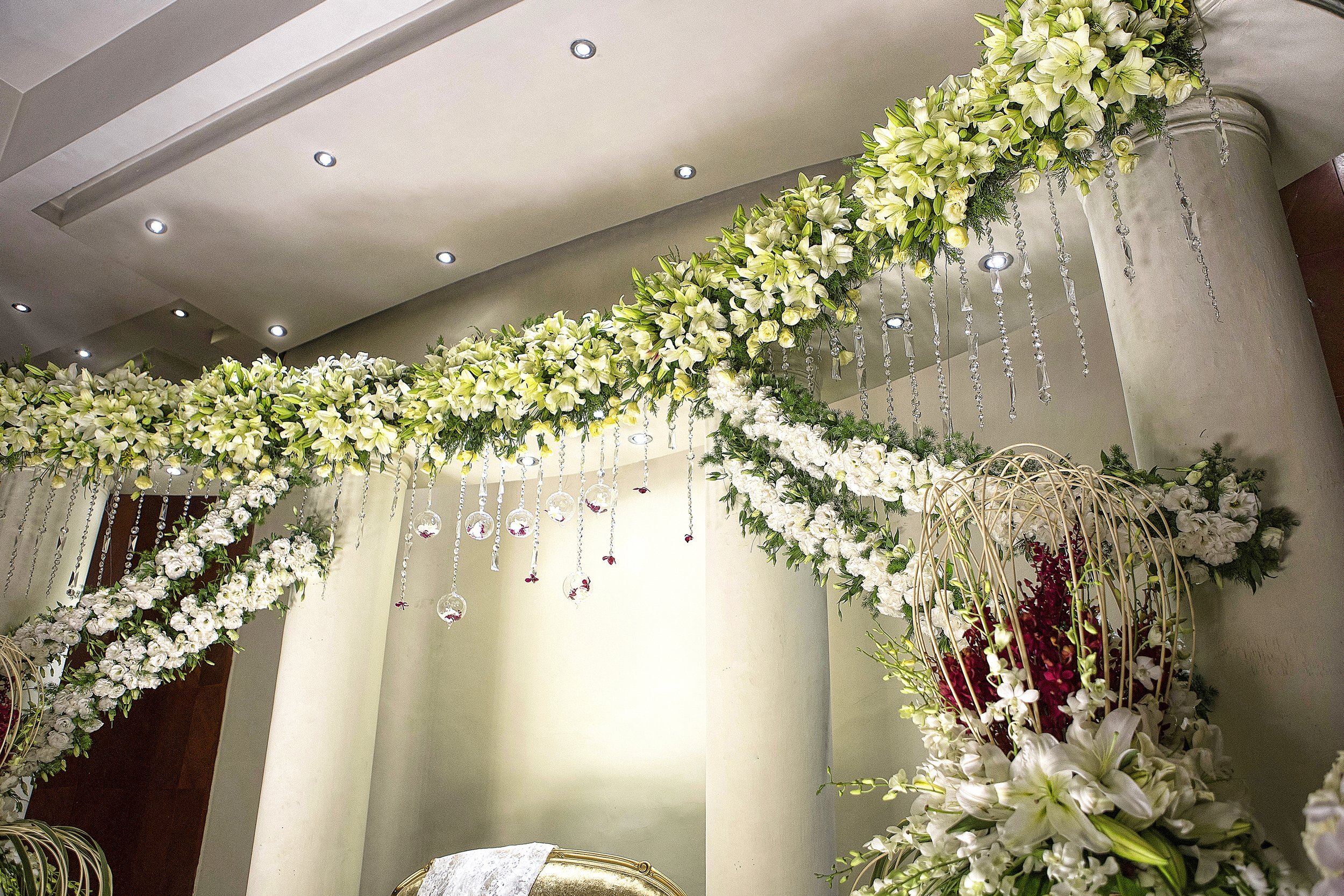 An elaborate indoor event floral display featuring large swags of white and green flowers (lilies, hydrangeas, orchids) draped across columns, with hanging crystal beads, clear glass baubles containing red accents, and a gold chair in the background.
