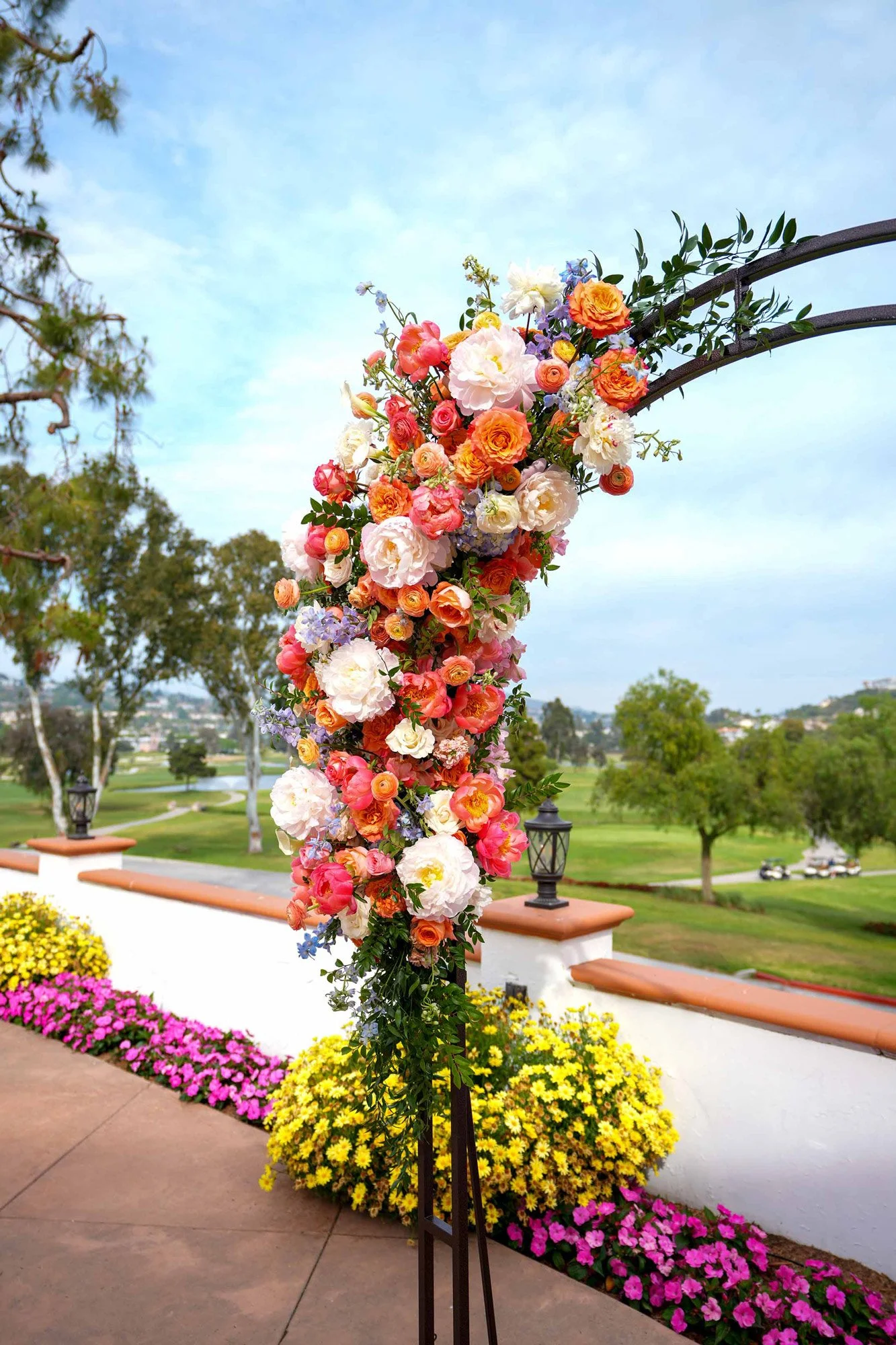 A black metal garden arch adorned with vibrant pink, orange, white, and blue flowers, set up outdoors on a patio overlooking a green golf course with rolling hills in the background.