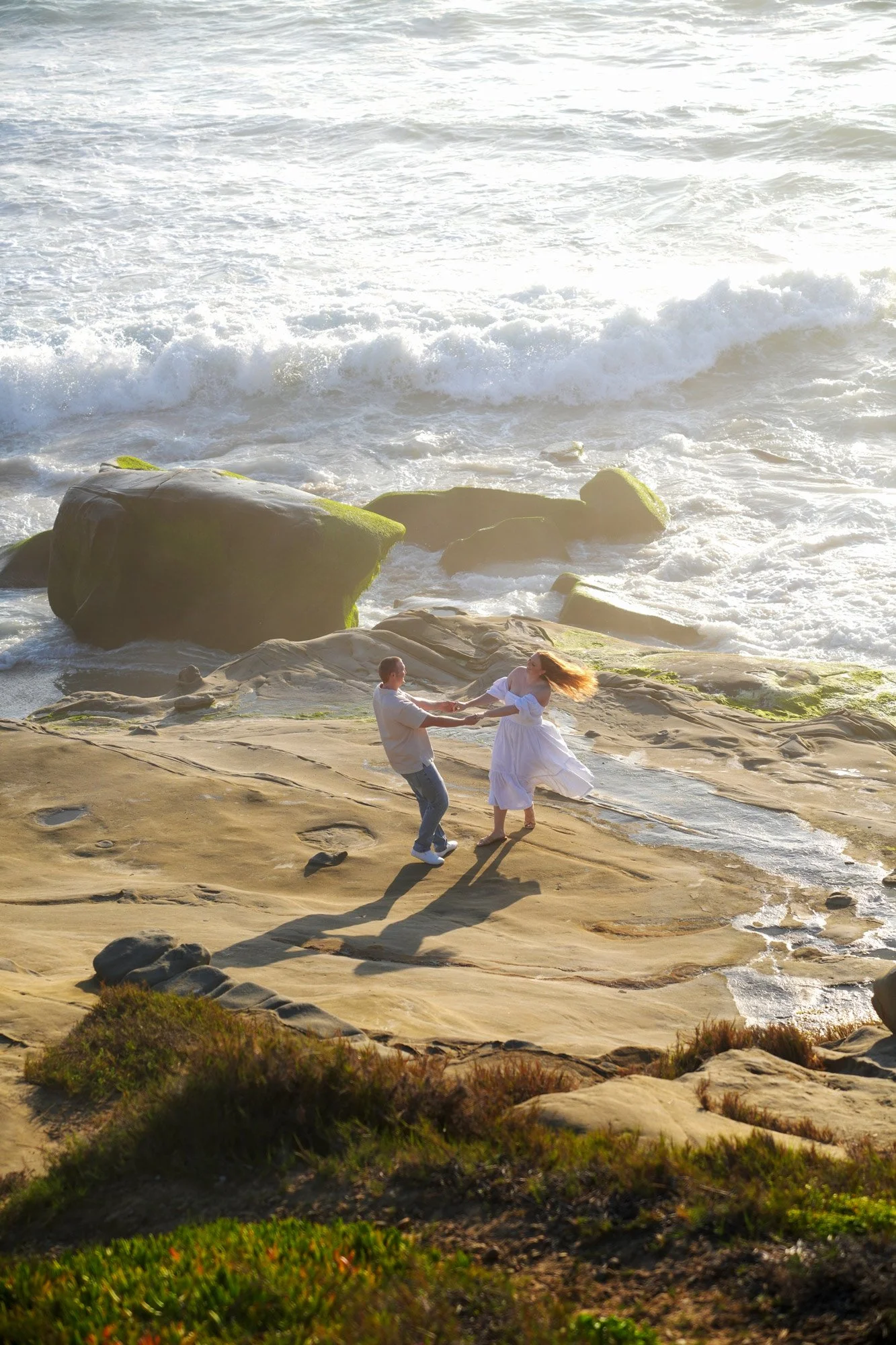 An outdoor photograph of a couple holding hands and spinning on a rocky beach near the ocean. The man wears a grey shirt and jeans, and the woman, in a white dress, has her red hair flowing with the motion. They are on tan-colored rock formations as 