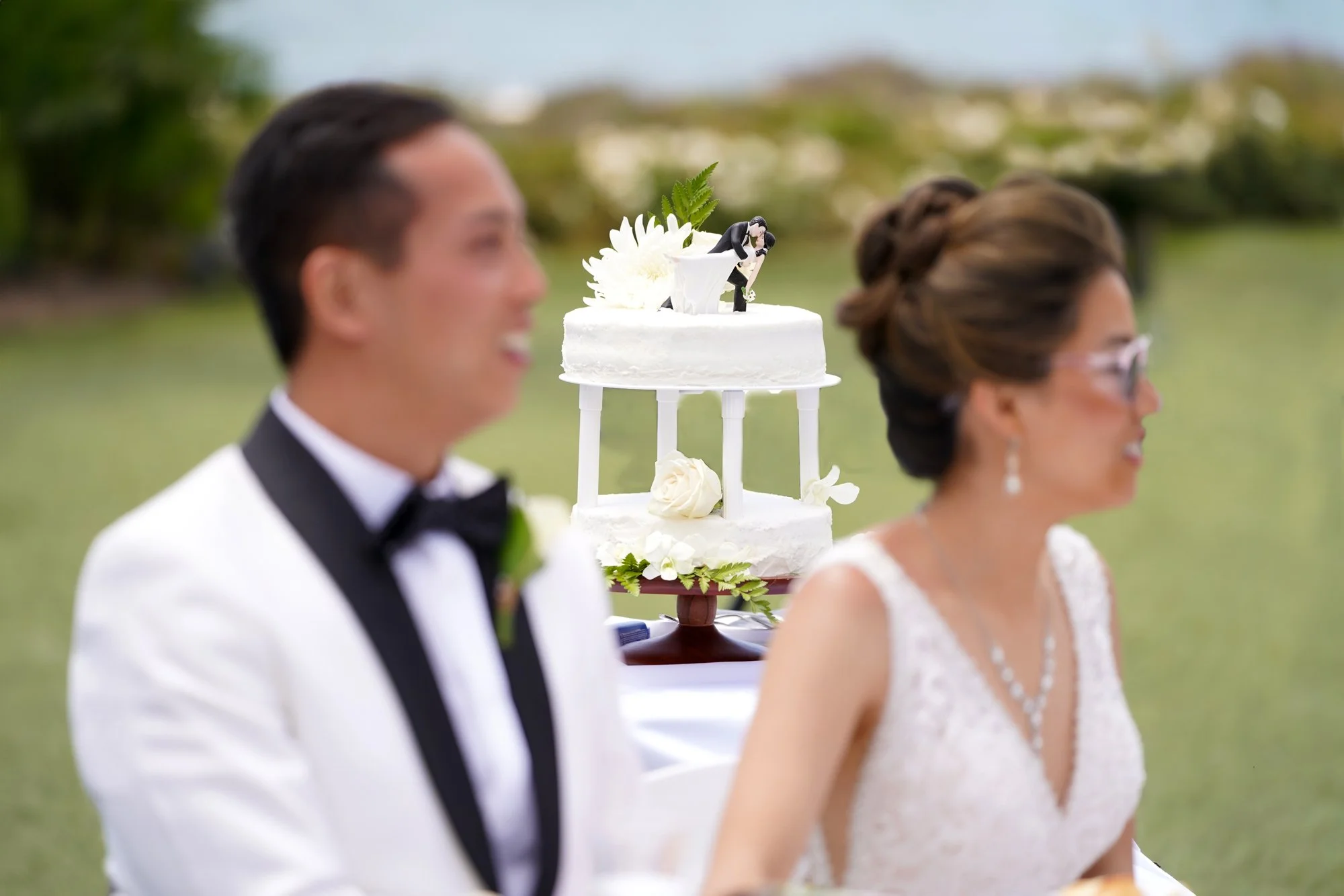 A bride in a white lace gown and a groom in a white tuxedo jacket sit smiling at their table, with a two-tiered wedding cake featuring a couple figurine topper between them during a sunny outdoor reception.