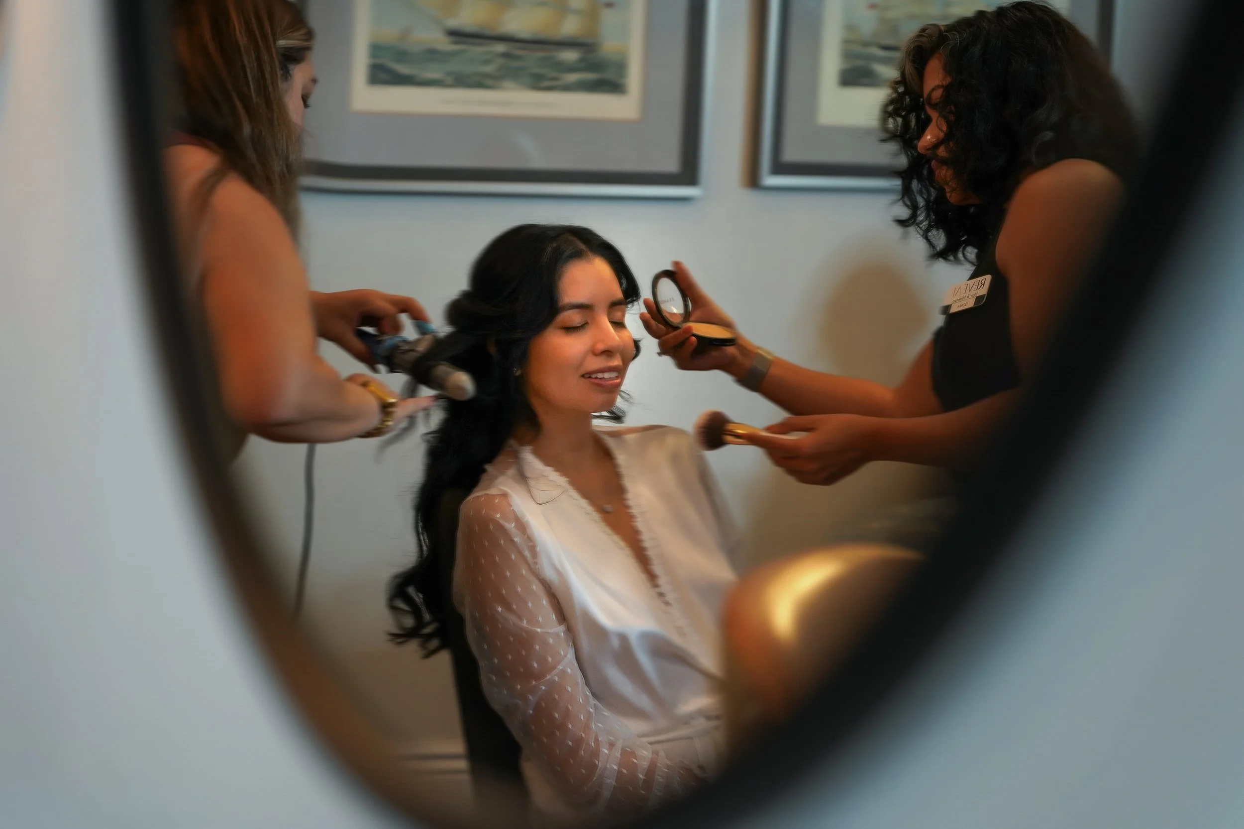 A bride sits for wedding day hair and makeup prep while wearing a white sheer lace-trimmed robe with long polka dot sleeves.