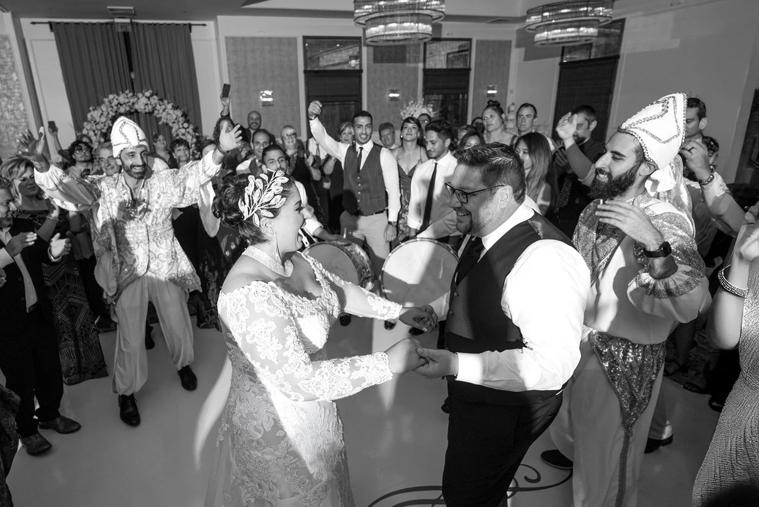 A black and white indoor wedding reception celebration. The bride, in a white lace dress, and the groom, in a black vest, are dancing and holding hands in the center of the image, smiling at each other. Guests surround them, including two men in orna