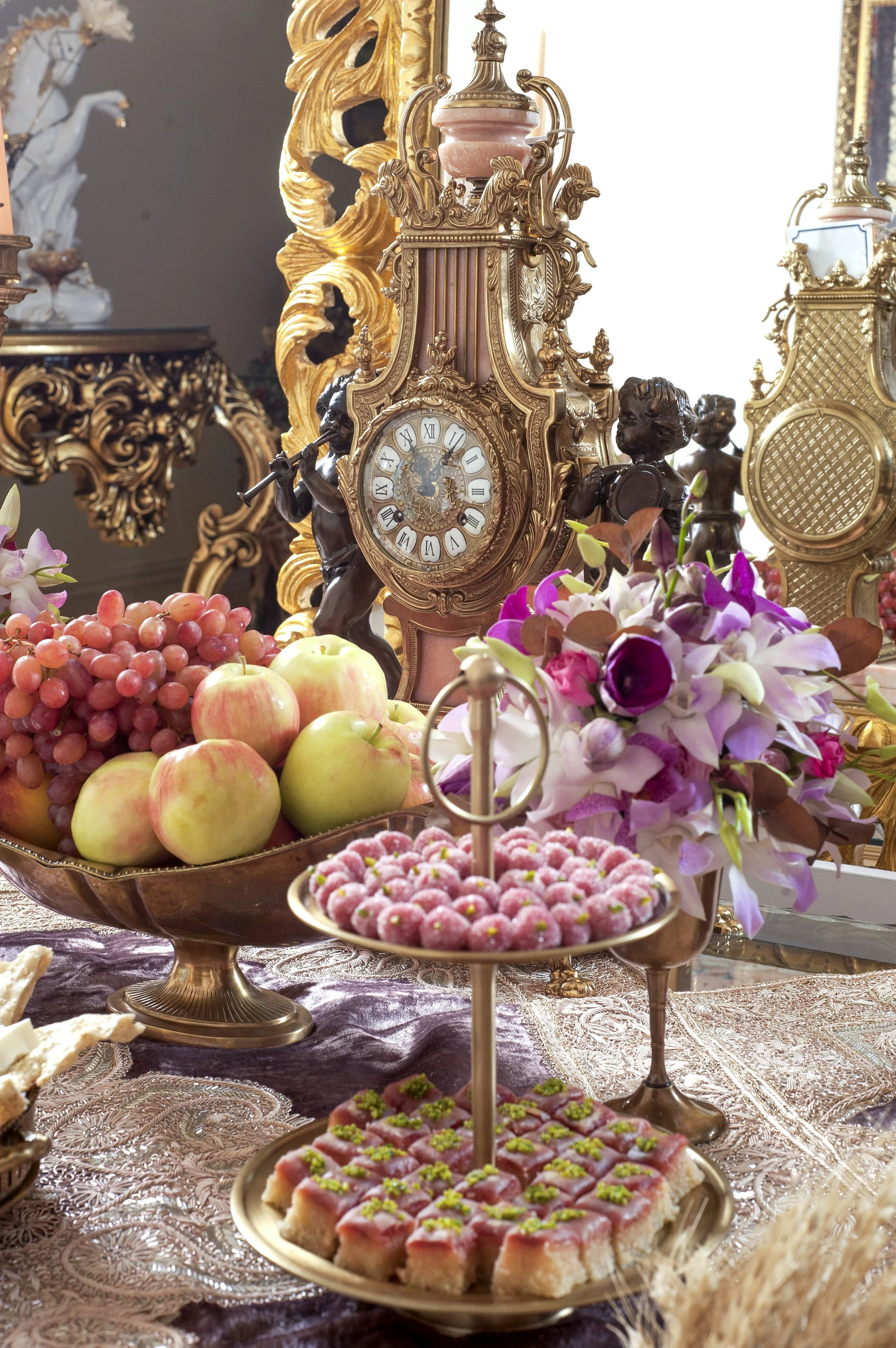 An elaborate, formal table setting, likely a Persian wedding Sofreh Aghd, featuring an antique gold clock, a two-tiered stand with pink and green sweets and pastries, a bowl of red grapes and green apples, white and purple floral arrangements, and or