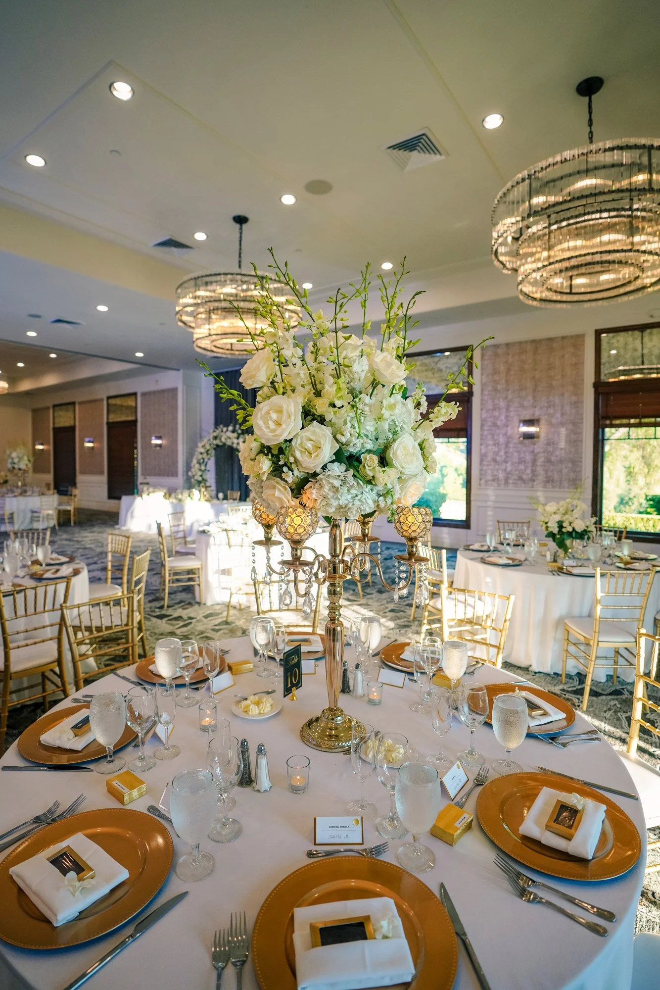 An indoor photograph of an elegantly set dining table for a formal event. The table features gold charger plates, white napkins, gold-rimmed glasses, and a prominent gold candelabra centerpiece with white floral arrangements. Other set tables, large 