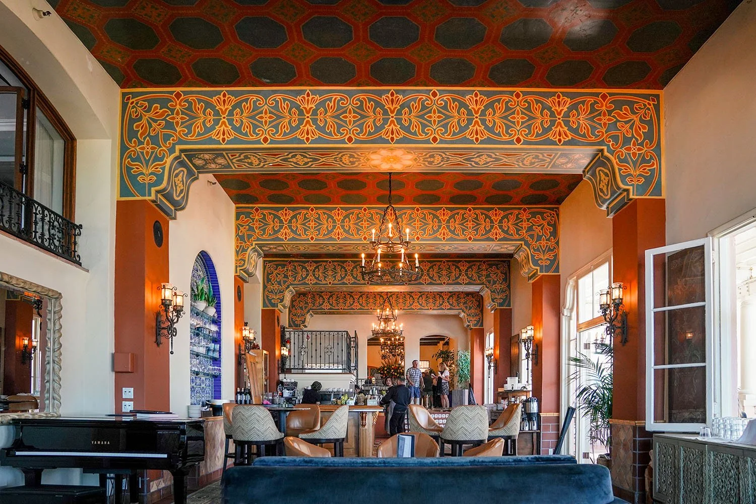 The opulent Spanish-inspired interior of the La Sala Lounge at the La Valencia Hotel in La Jolla, California, features a soaring, hand-painted cathedral-style ceiling with gold accents and orange/teal patterns, dark wood furniture, and a bar area.