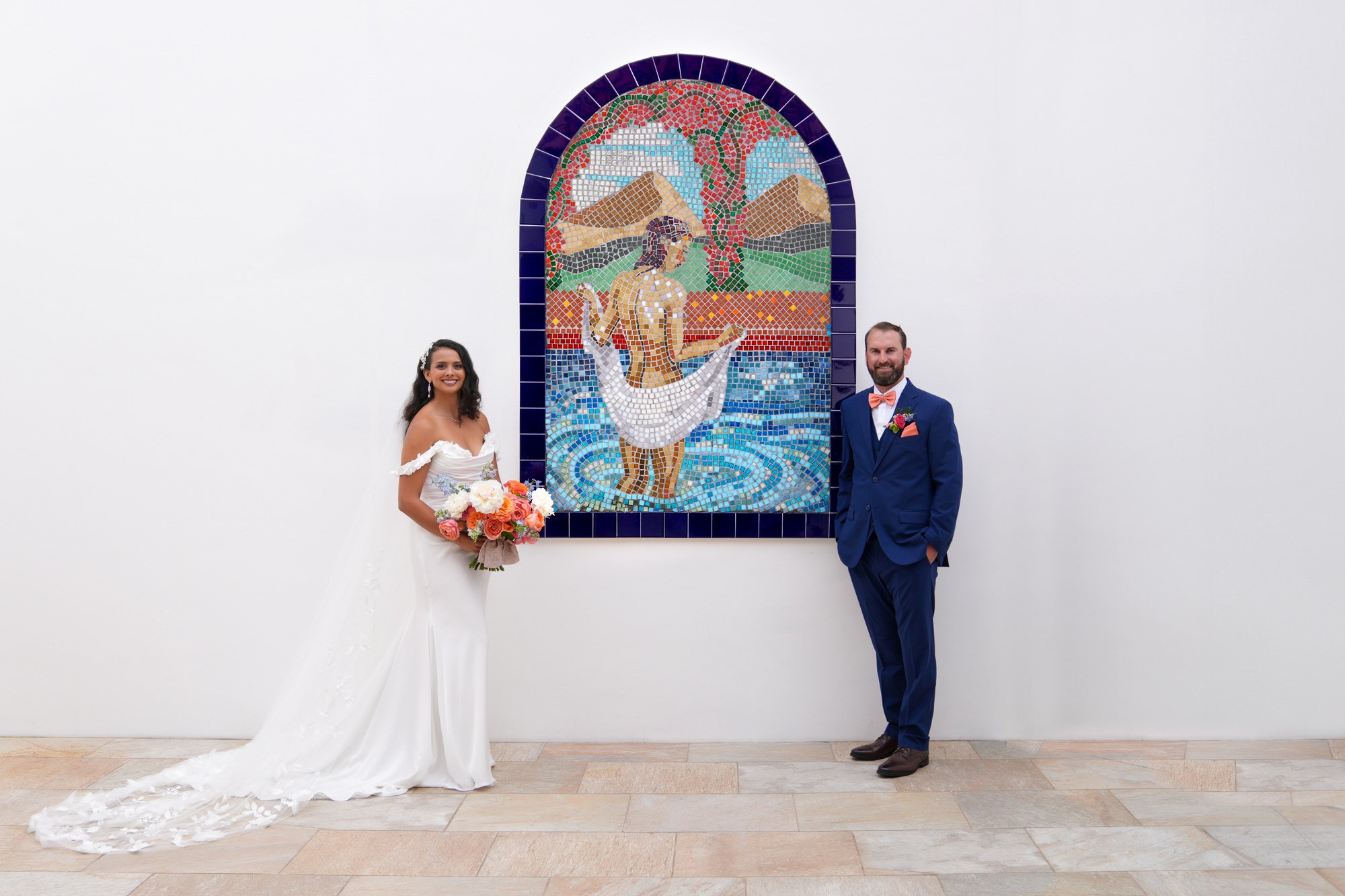 A bride in a white, off-the-shoulder gown and a groom in a blue suit and orange bowtie stand on a tiled patio beside a large, arched mosaic artwork depicting a woman in water in La Costa Resort.


