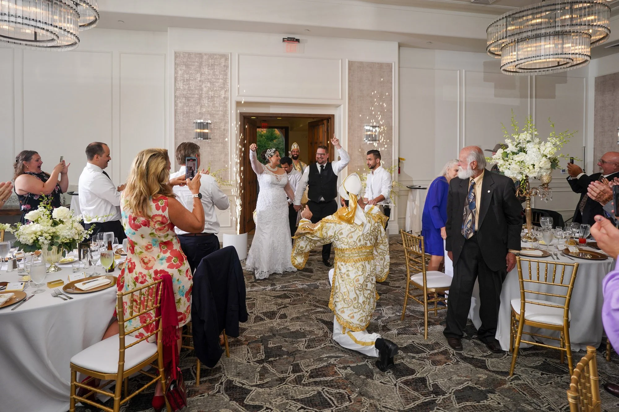 An indoor photograph capturing the enthusiastic entry of a newly married couple into their wedding reception at The Maderas Golf Club. The bride wears a white wedding dress, and the groom wears a black vest and white shirt. They enter through an arch
