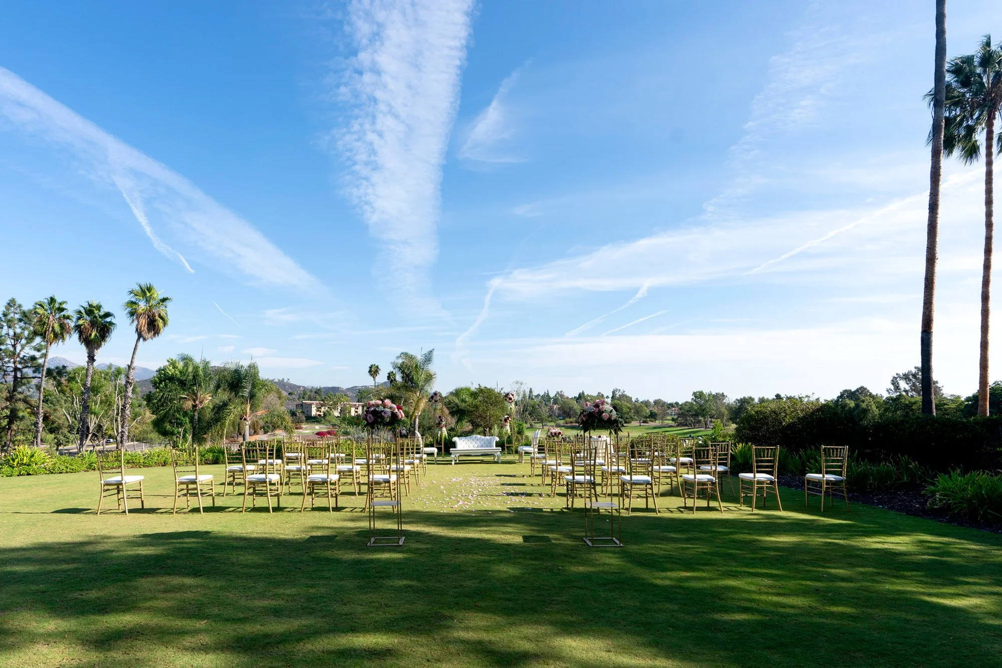 A wide, outdoor photograph of a wedding ceremony set up on a grassy lawn. Rows of gold chairs with white cushions face an altar area with a white couch or bench. The scene is set under a bright blue sky with white clouds and is surrounded by lush gre