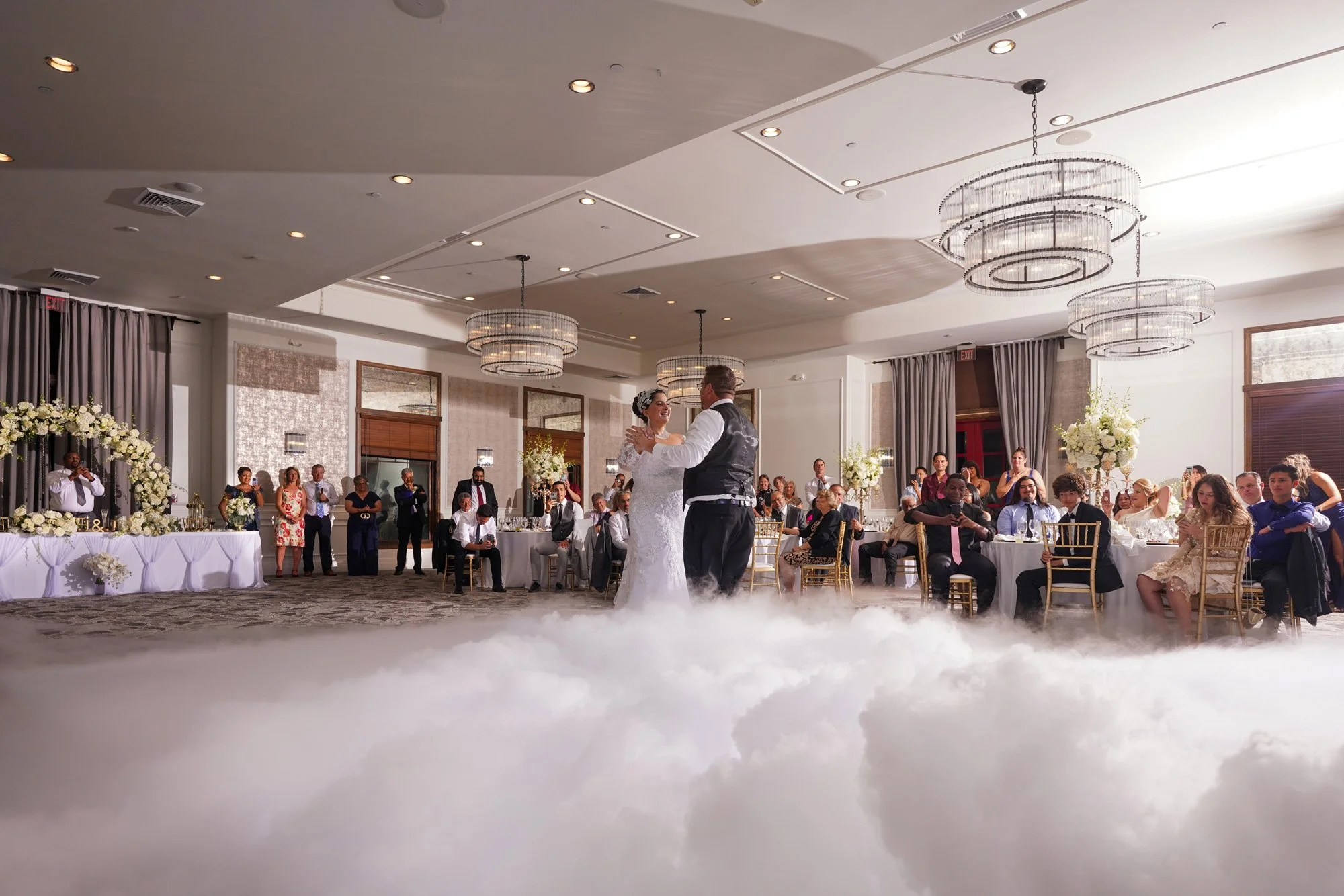 An indoor photograph capturing a wedding reception featuring the bride and groom dancing their first dance on a dance floor covered in a thick layer of white dry ice fog. The bride wears a white dress, and the groom wears a black vest over a white sh